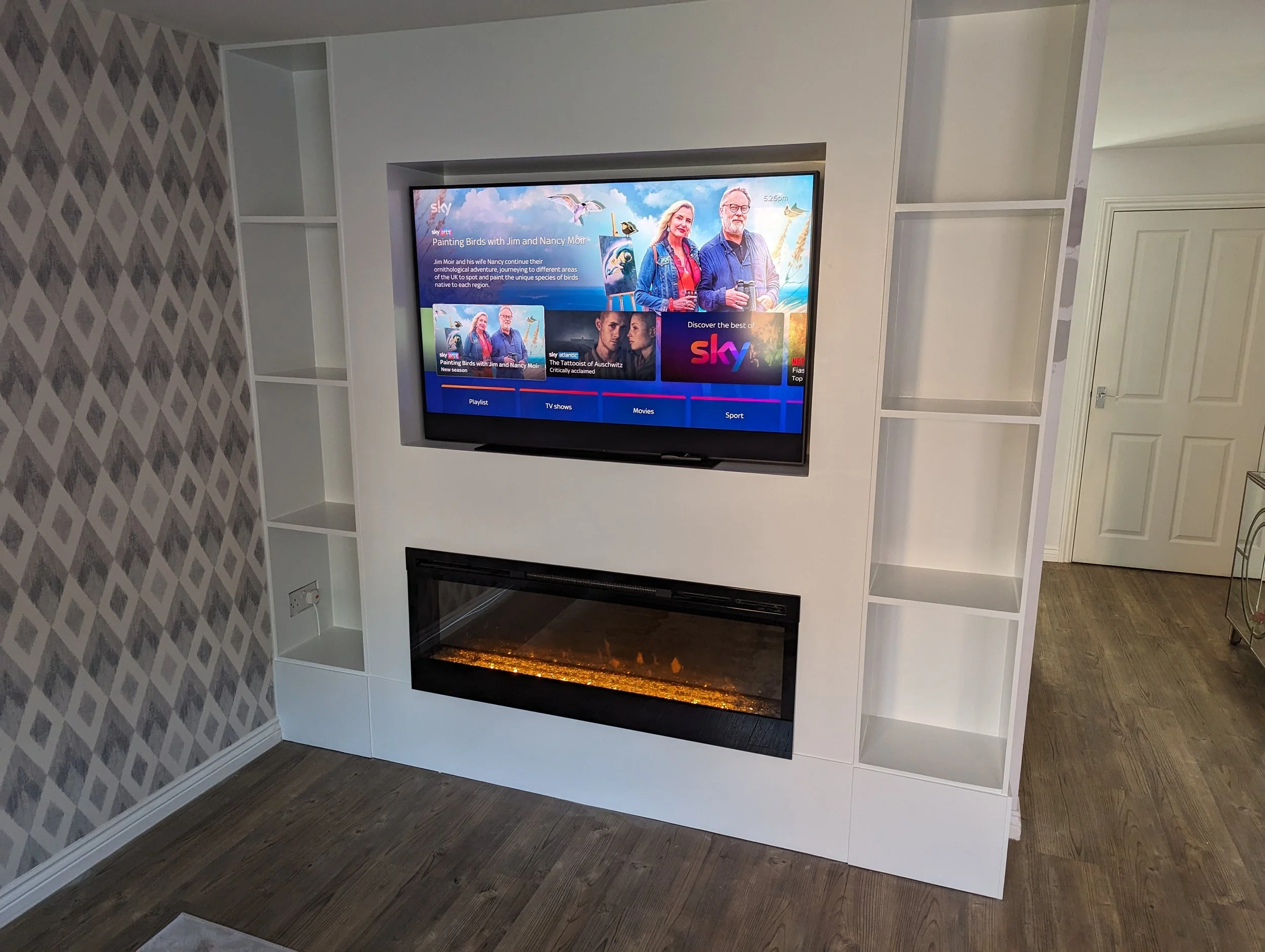Living room entertainment wall with a mounted flat-screen TV displaying the Sky TV interface and a built-in electric fireplace below it. The wall has white shelves on either side of the TV, with a patterned wallpaper on the left side and a door on th
