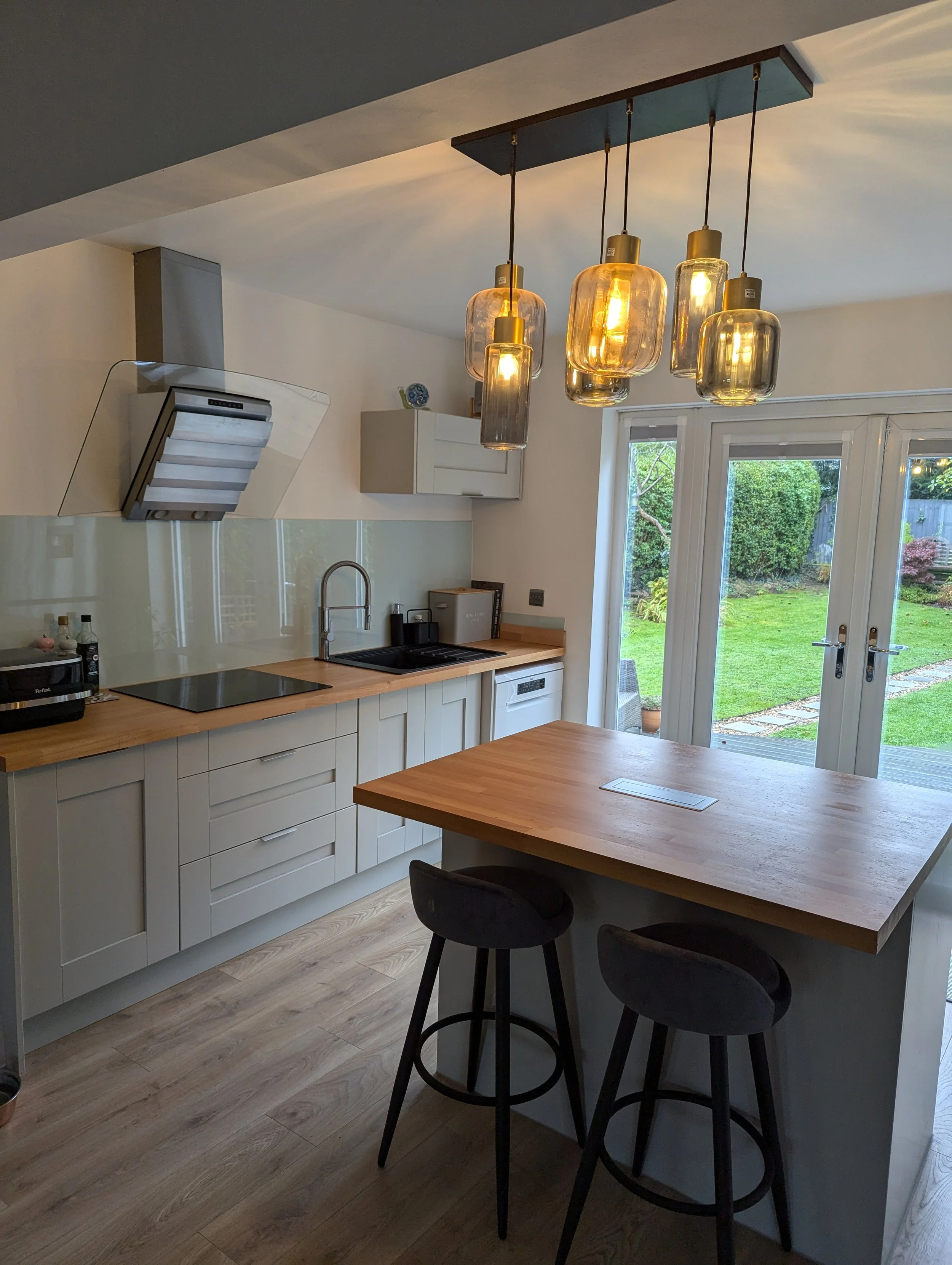 Modern kitchen with white cabinetry, wooden countertops, and a wooden dining island with two black chairs. Pendant lights hang above the island. There are glass doors leading to a backyard with grass and stone pathway.