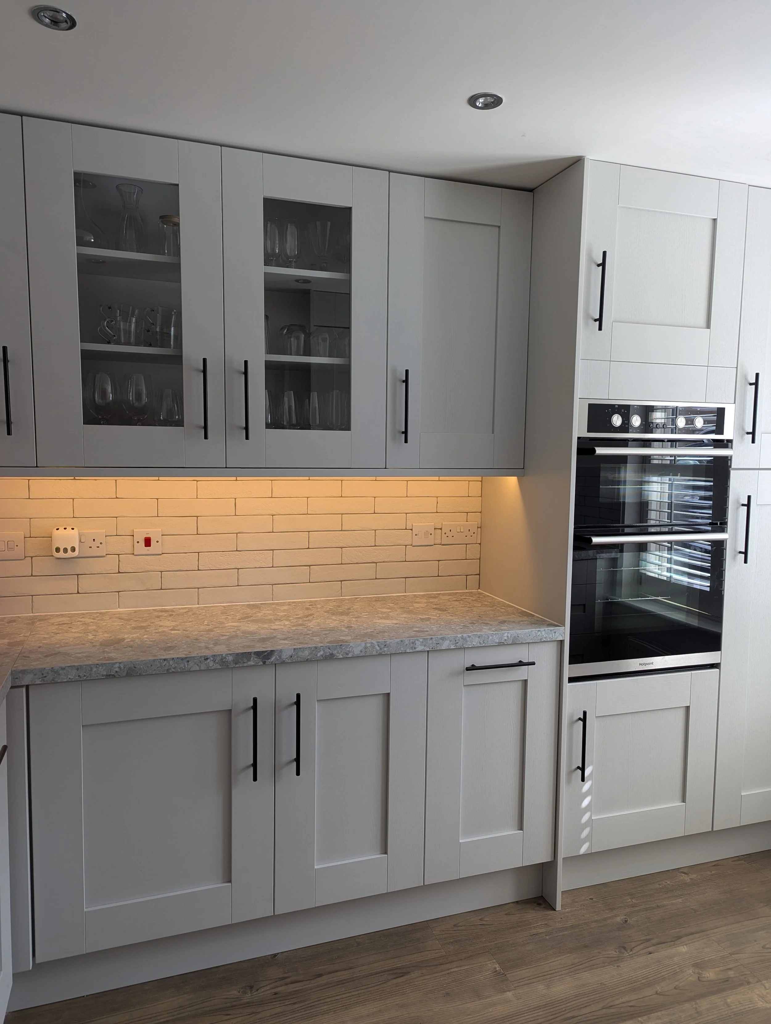 Modern kitchen with light gray cabinets, glass-paneled upper cabinets filled with glassware, a marble countertop, brick backsplash, and built-in double oven.