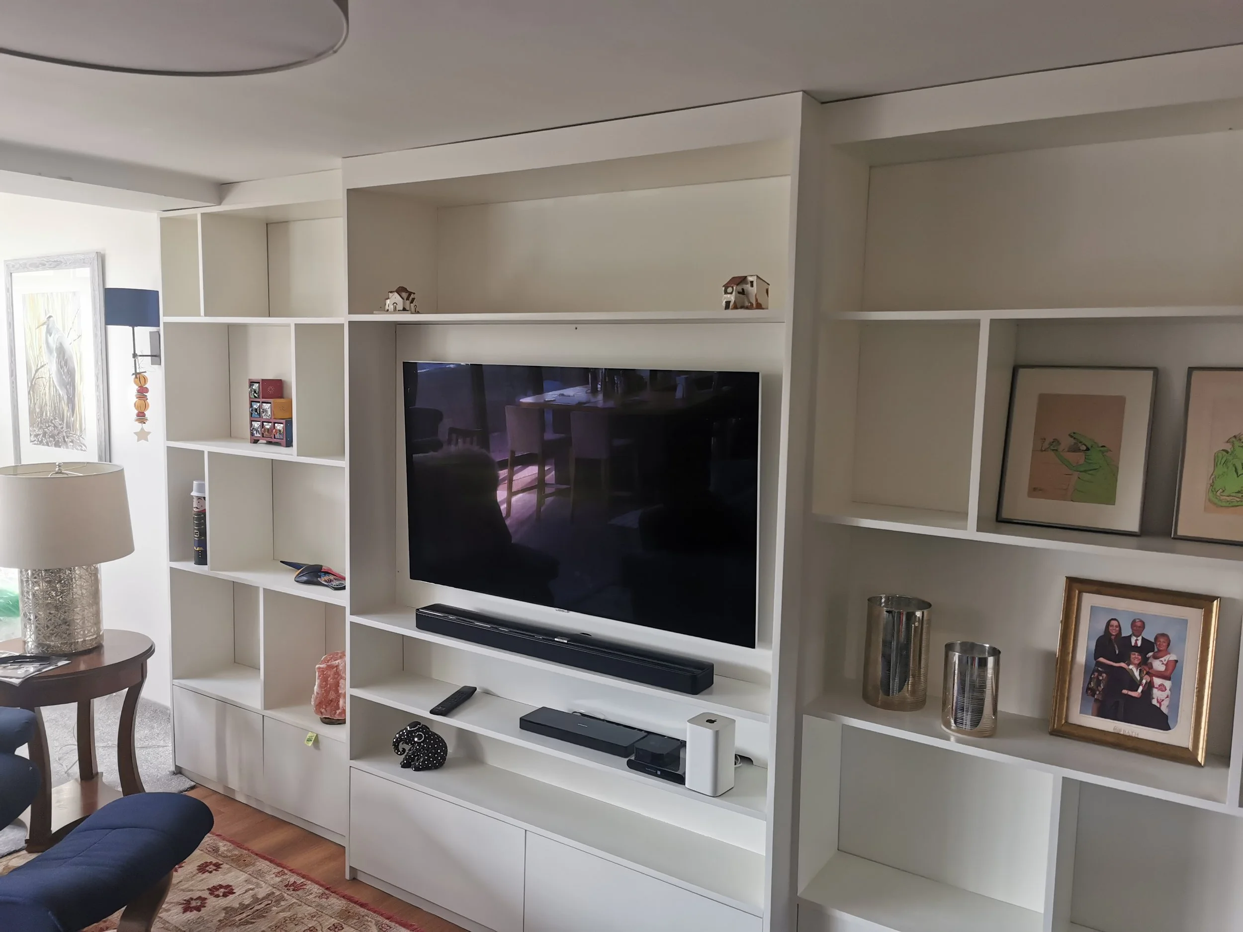 Living room with white entertainment center holding a wall-mounted flat-screen TV, music soundbar, and various decorative items including framed pictures, vases, and a salt lamp, with a window and artwork on the wall.