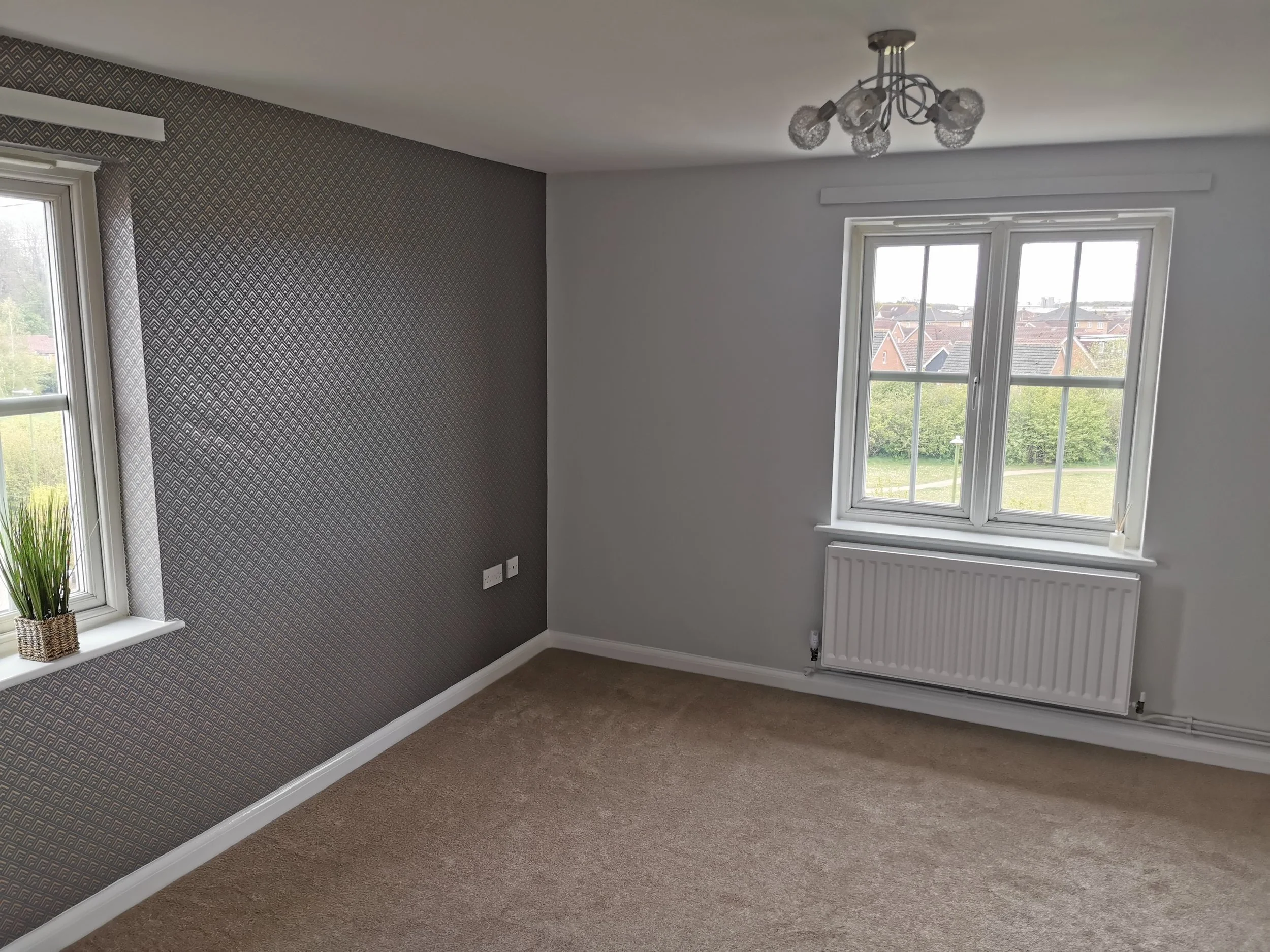 Empty living room with beige carpet, white walls, large windows, one with patterned wallpaper, a potted plant on the windowsill, a radiator under one window, and a ceiling light fixture.
