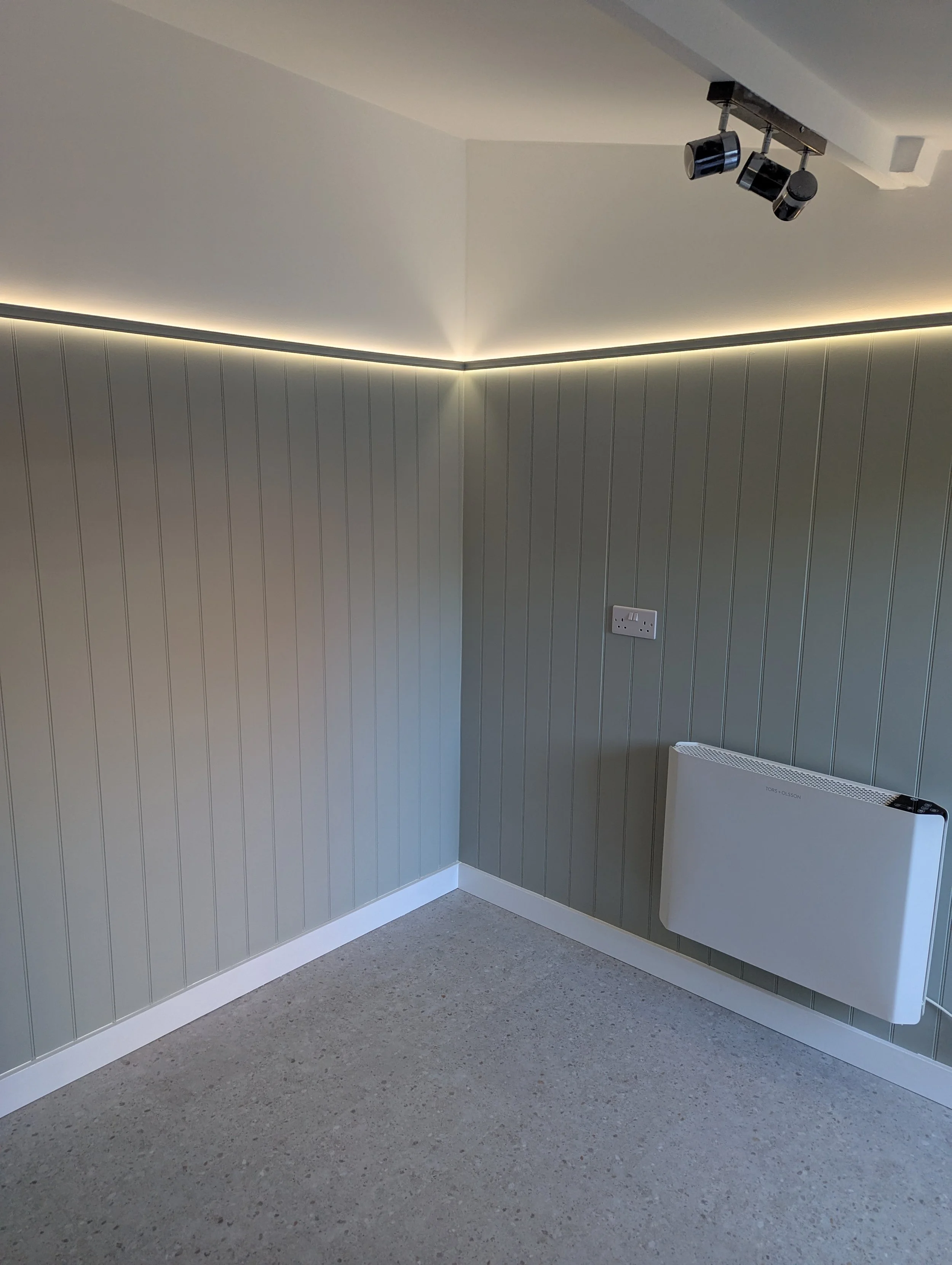 Empty room with textured gray walls, a white baseboard, a wall-mounted heater, a double power outlet, and ceiling spotlights with track lighting.