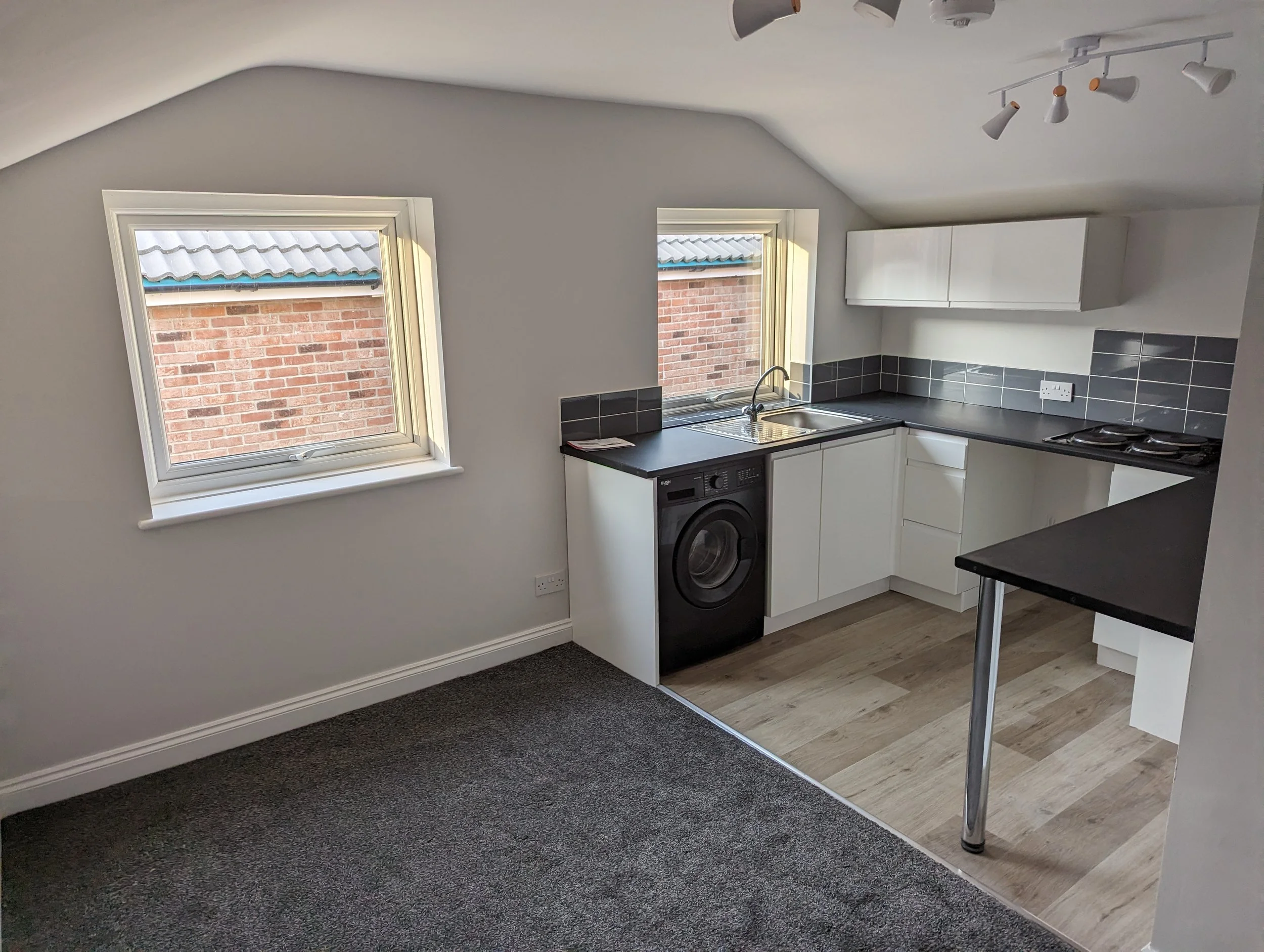 Small kitchen with two windows, black countertop, white cabinets, gray tile backsplash, washing machine, sink, stove, and overhead light fixtures.