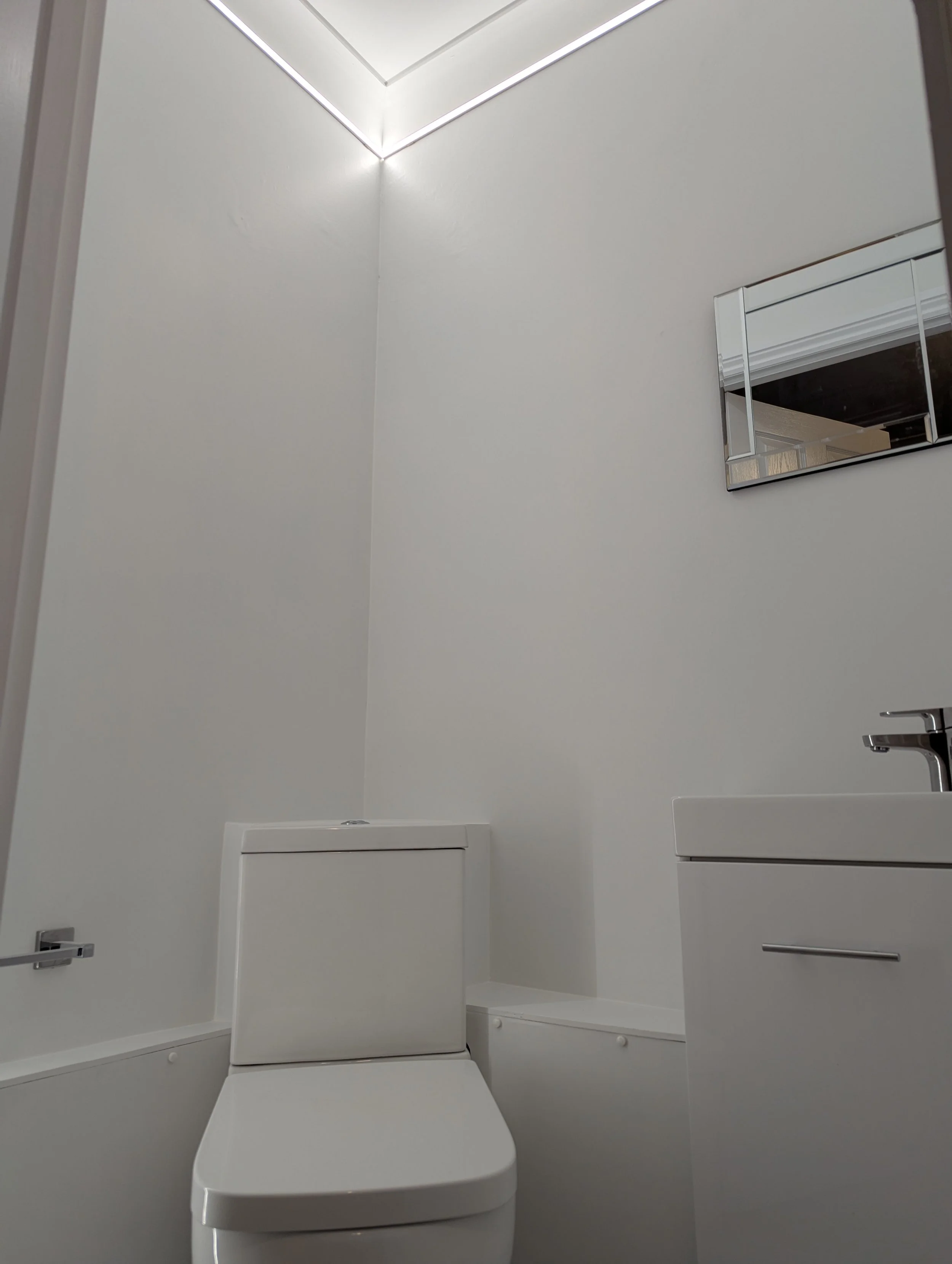 Minimalist white bathroom with a toilet, small white sink, and a rectangular mirror on the wall.