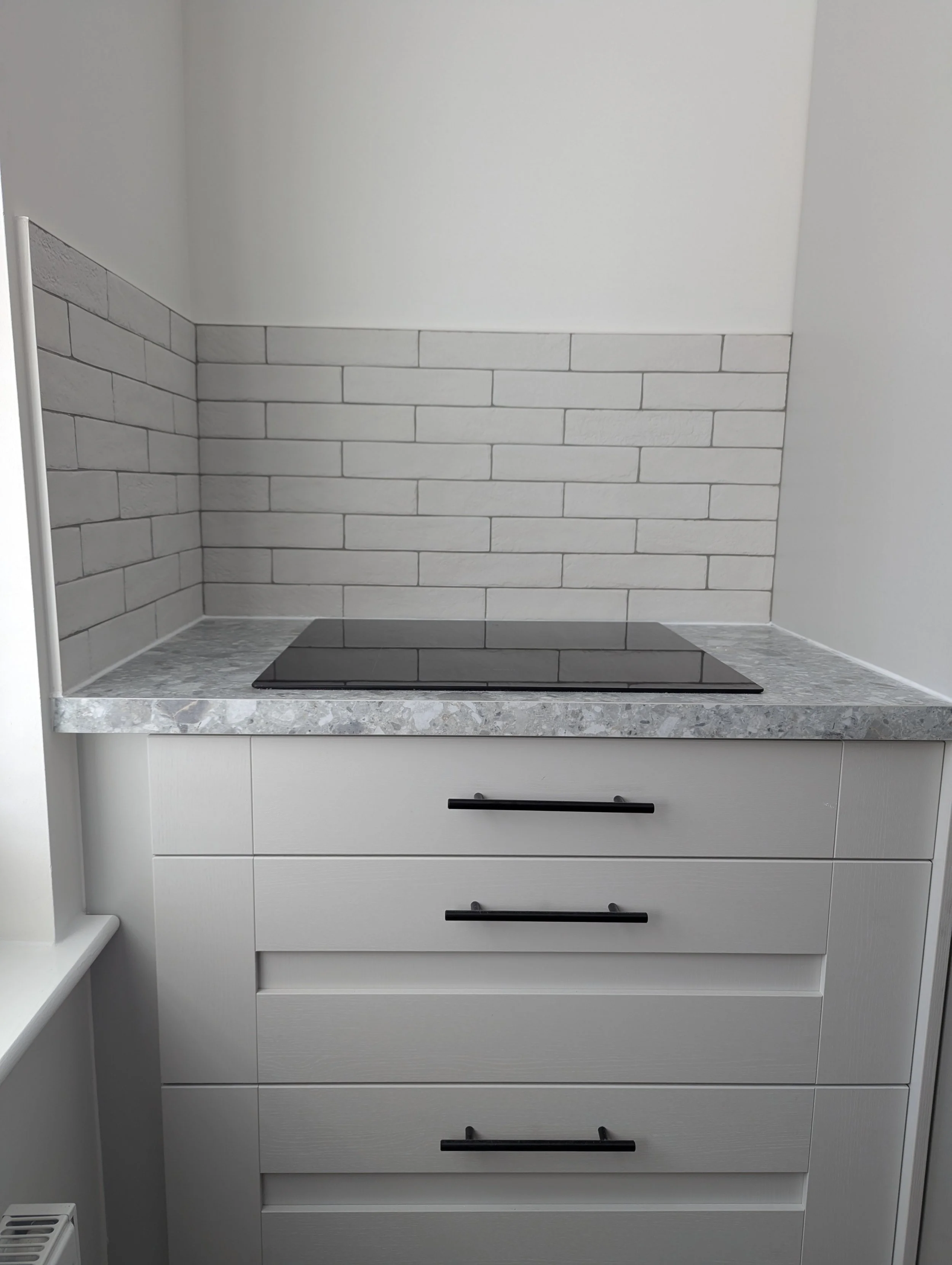 Kitchen countertop with stovetop, white cabinets, gray towel bar, tiled backsplash, and a small window on the left.