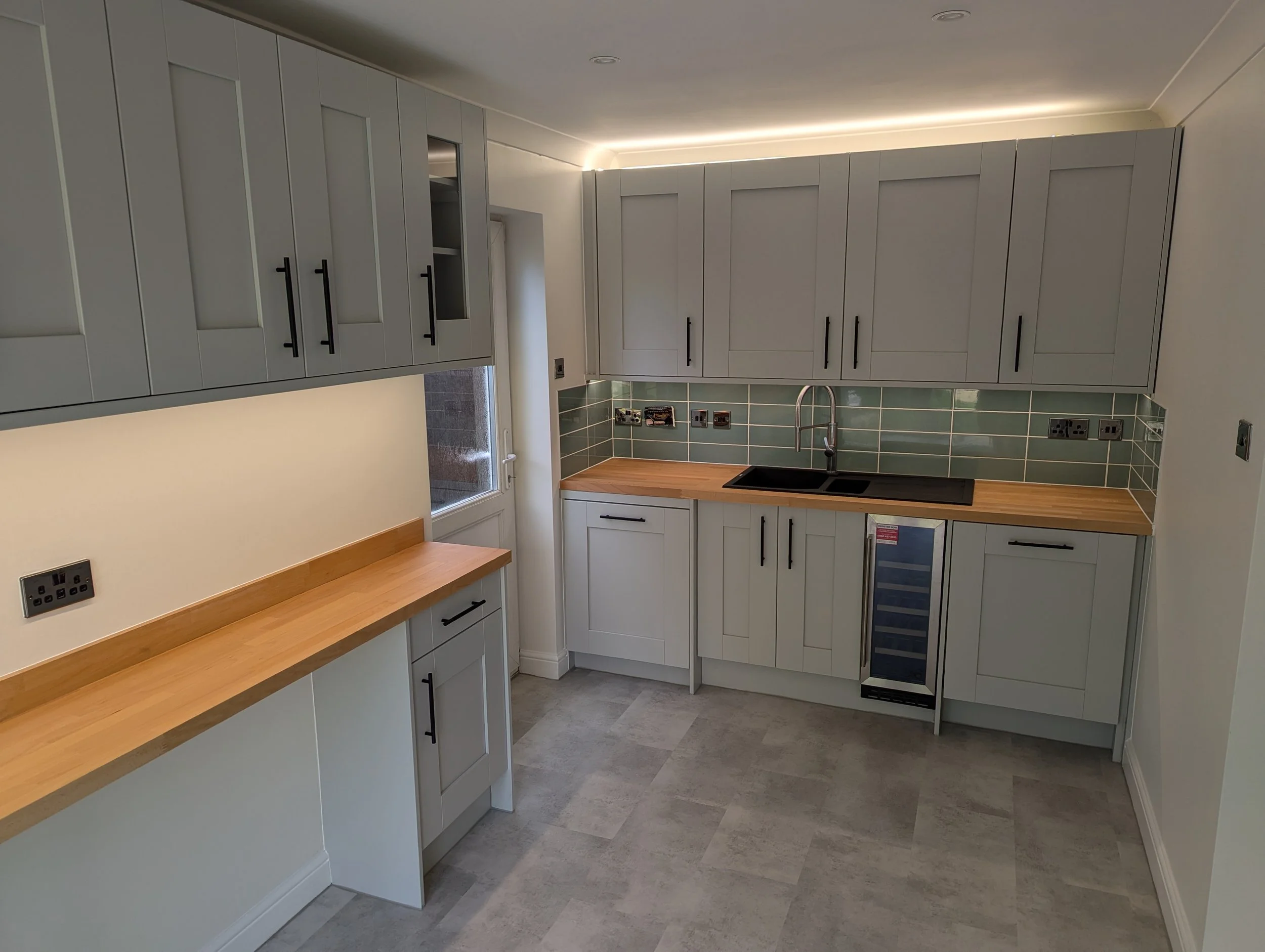 A modern kitchen with light gray cabinets, a black sink, a wooden countertop, gray tiles on the backsplash, a small wine cooler, and electrical outlets on the walls.