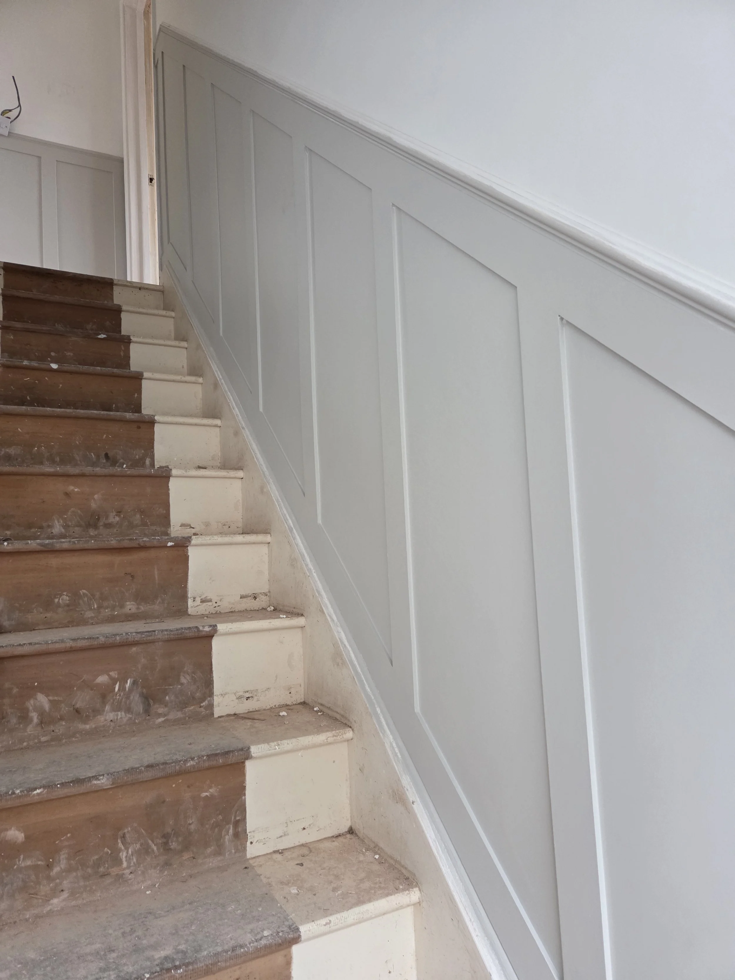 Partially renovated staircase with unfinished wooden steps and newly installed white wainscoting on the wall.