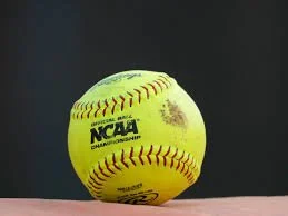 A yellow NCAA softball with red stitching and some dirt marks on a dark background.