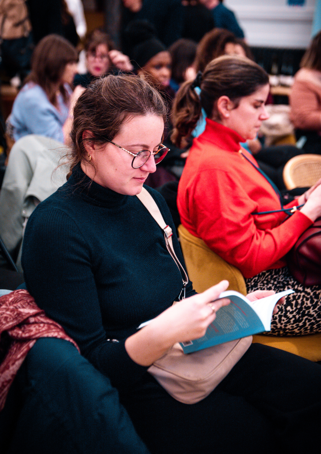 Plusieurs femmes assises en salle, certaines lisant des livres, dans une ambiance d'événement ou de conférence.