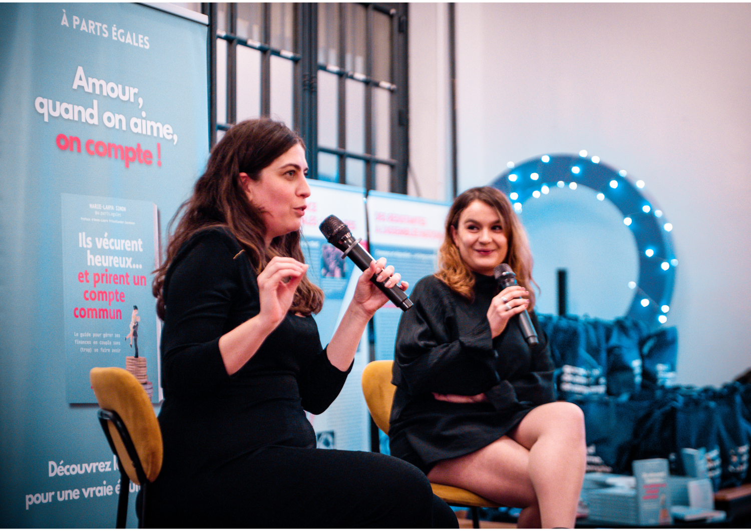 Deux femmes parlent lors d'une conférence, l'une avec un micro, devant un panneau publicitaire bleu avec du texte en français, et un décor lumineux avec un cercle de lumières bleues en arrière-plan.