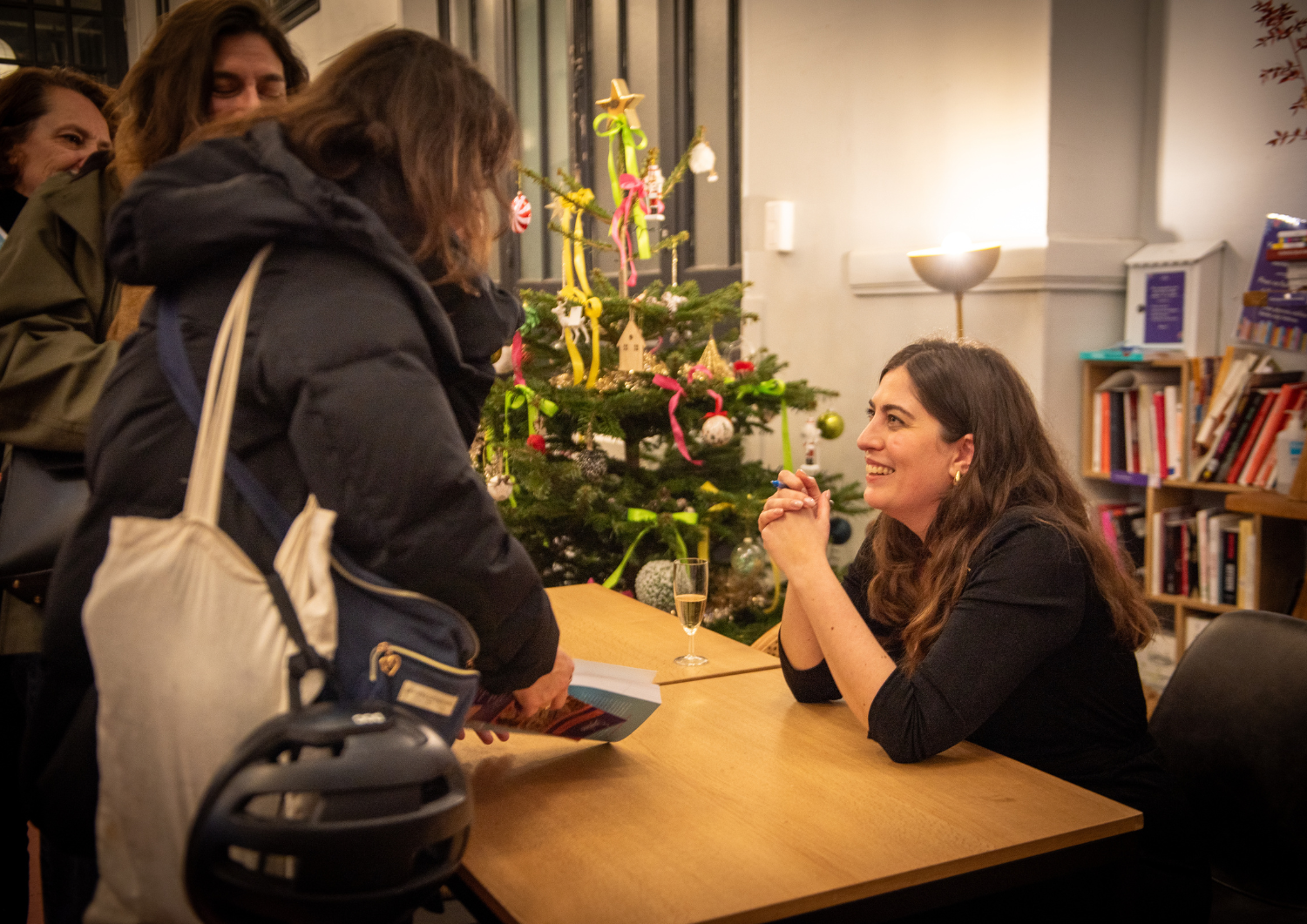 Une femme souriante, assise à une table, échange avec une autre femme qui lui tend un livre. Il y a un sapin de Noël décoré en arrière-plan, dans une salle avec des livres sur une étagère.