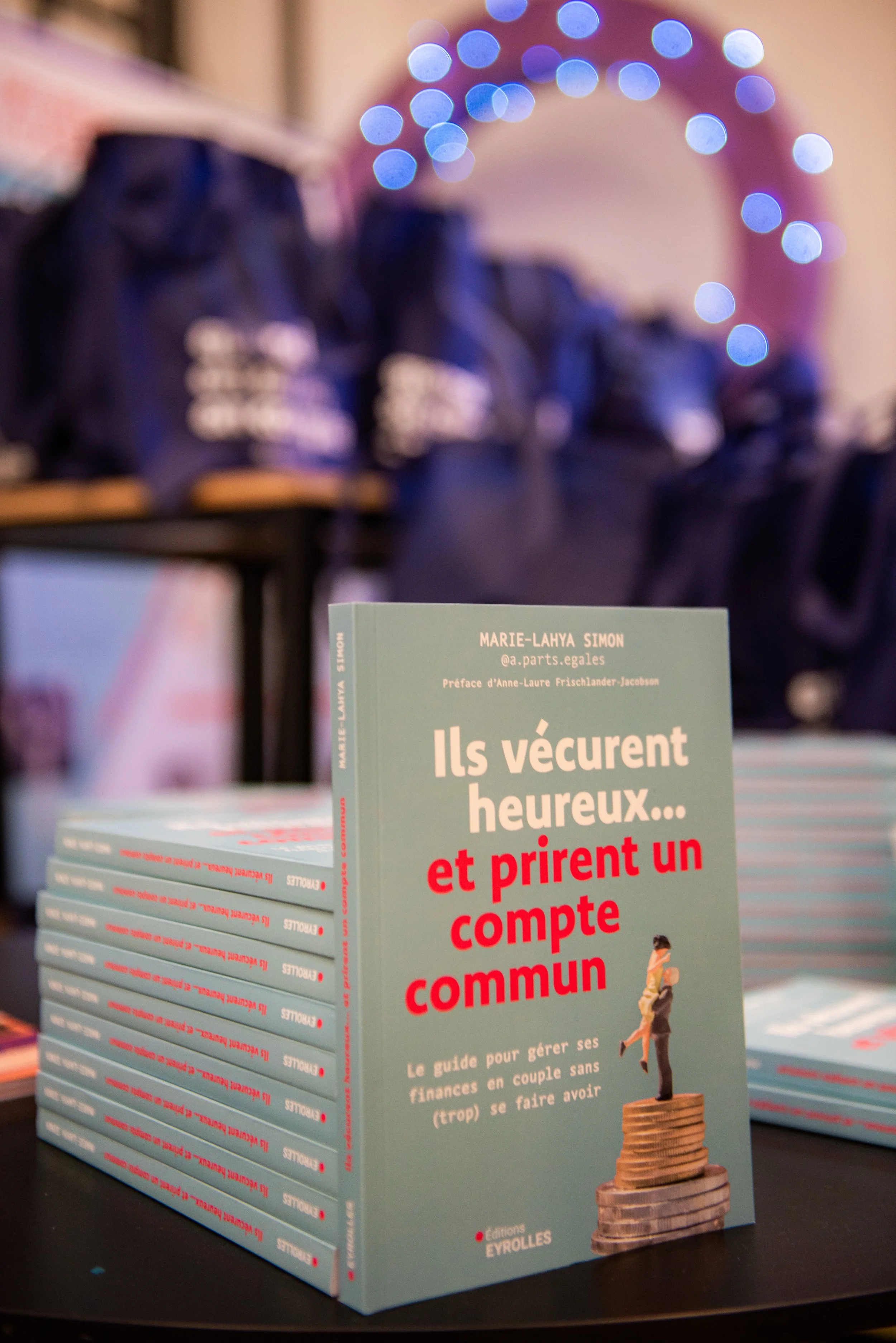 Couverture d'un livre intitulé "Ils vécurent heureux... et prient un compte commun", écrit par Marie-Lahya Simon, affiché sur une table avec d'autres copies empilées, dans un environnement avec des décorations lumineuses floues en arrière-plan.