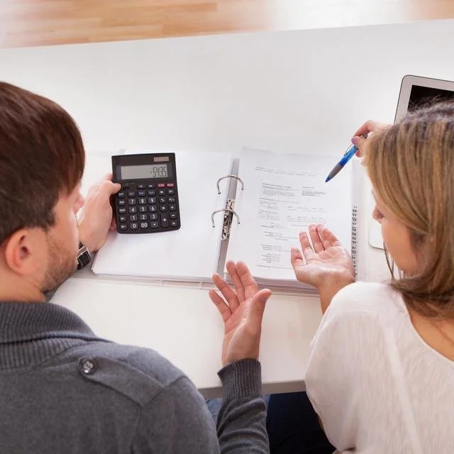 Deux personnes discutent, avec une calculatrice, des documents et une tablette sur une table blanche.