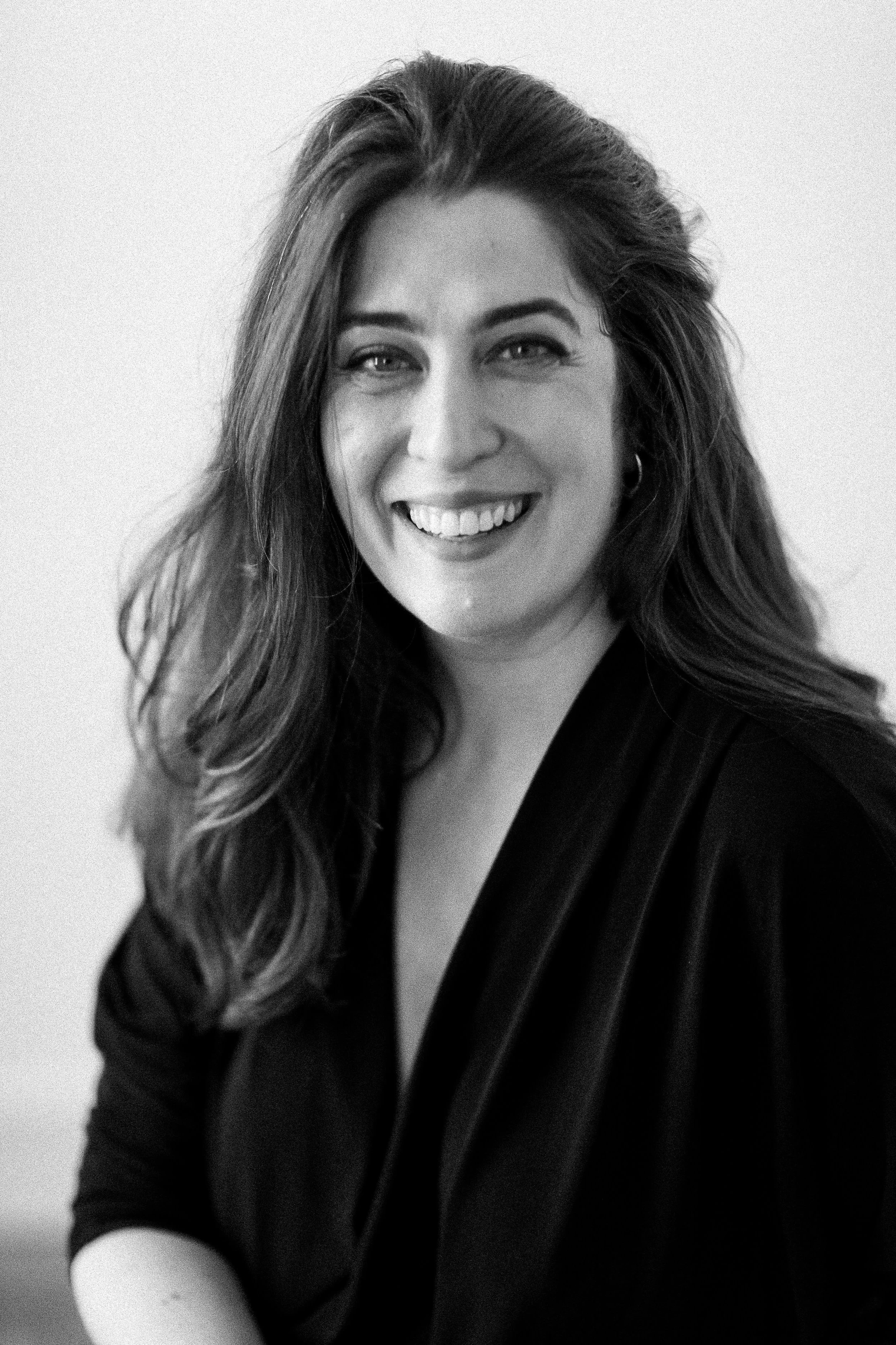Portrait en noir et blanc d'une femme souriante avec de longs cheveux ondulés, portant une veste noire.