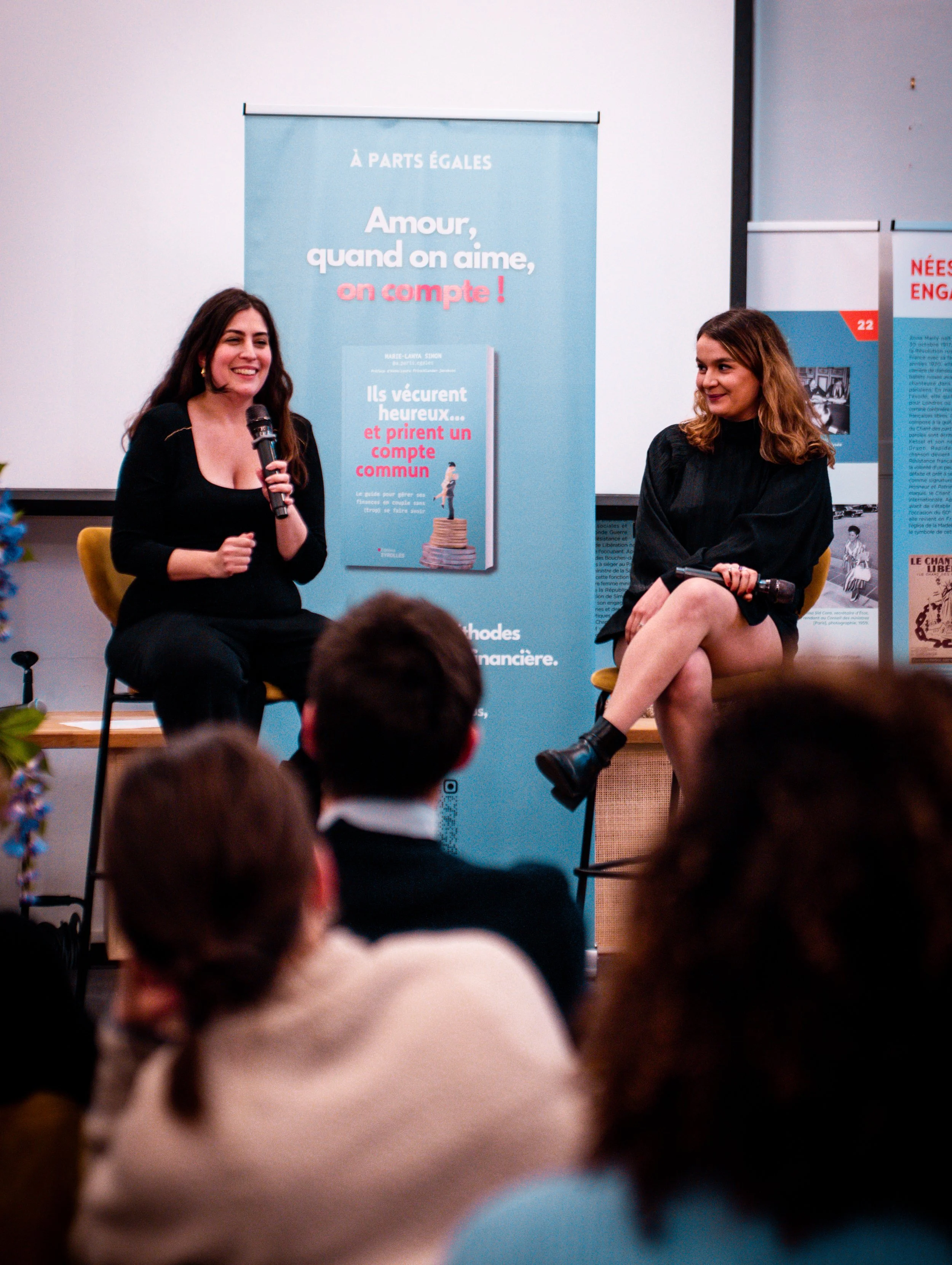 Deux femmes assises sur scène lors d'une discussion, avec une audience en face. Un panneau bleu derrière elles affiche un livre et un message promotionnel.