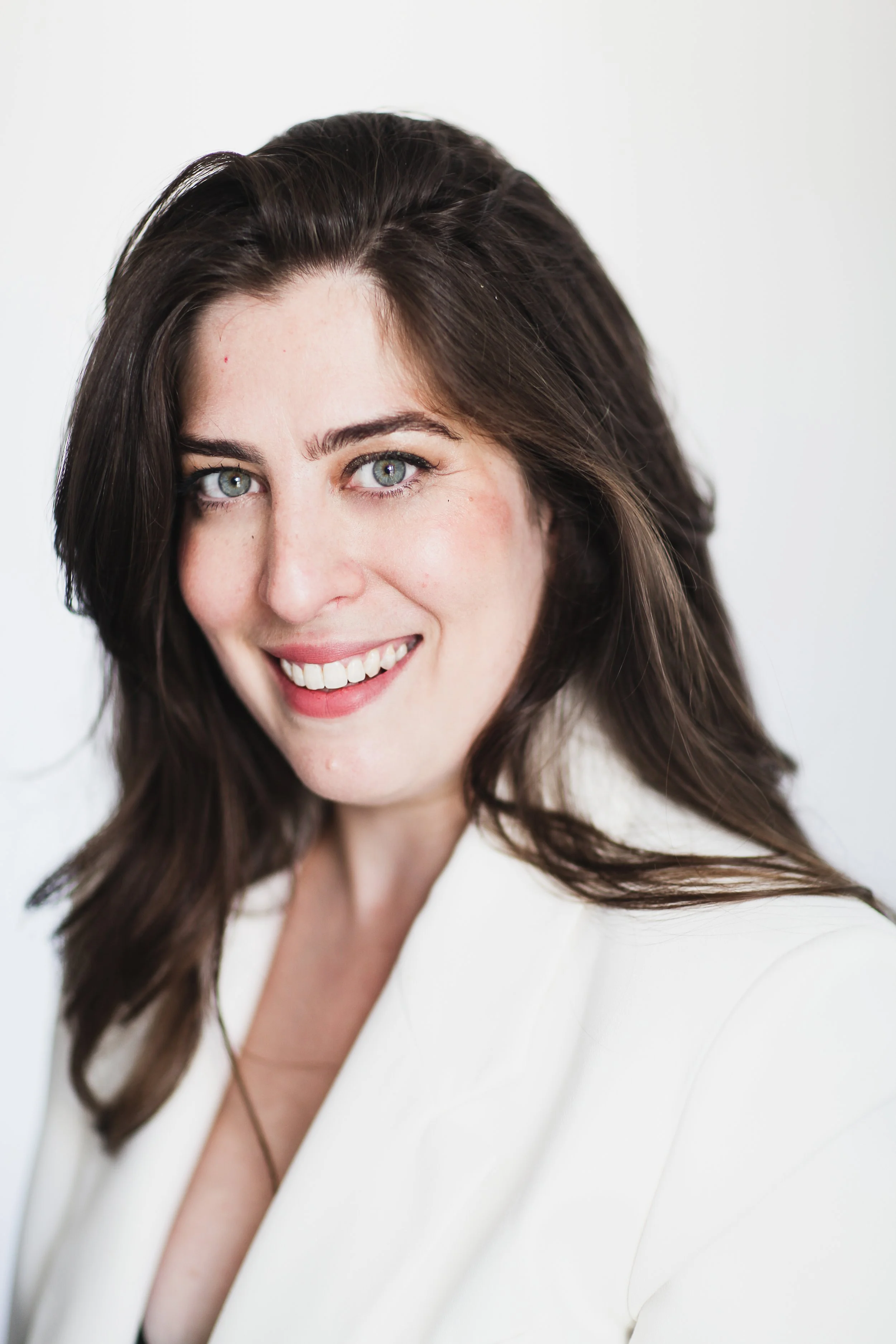 Une femme avec des cheveux bruns, souriante, portant une veste blanche, photographiée en gros plan contre un fond blanc.