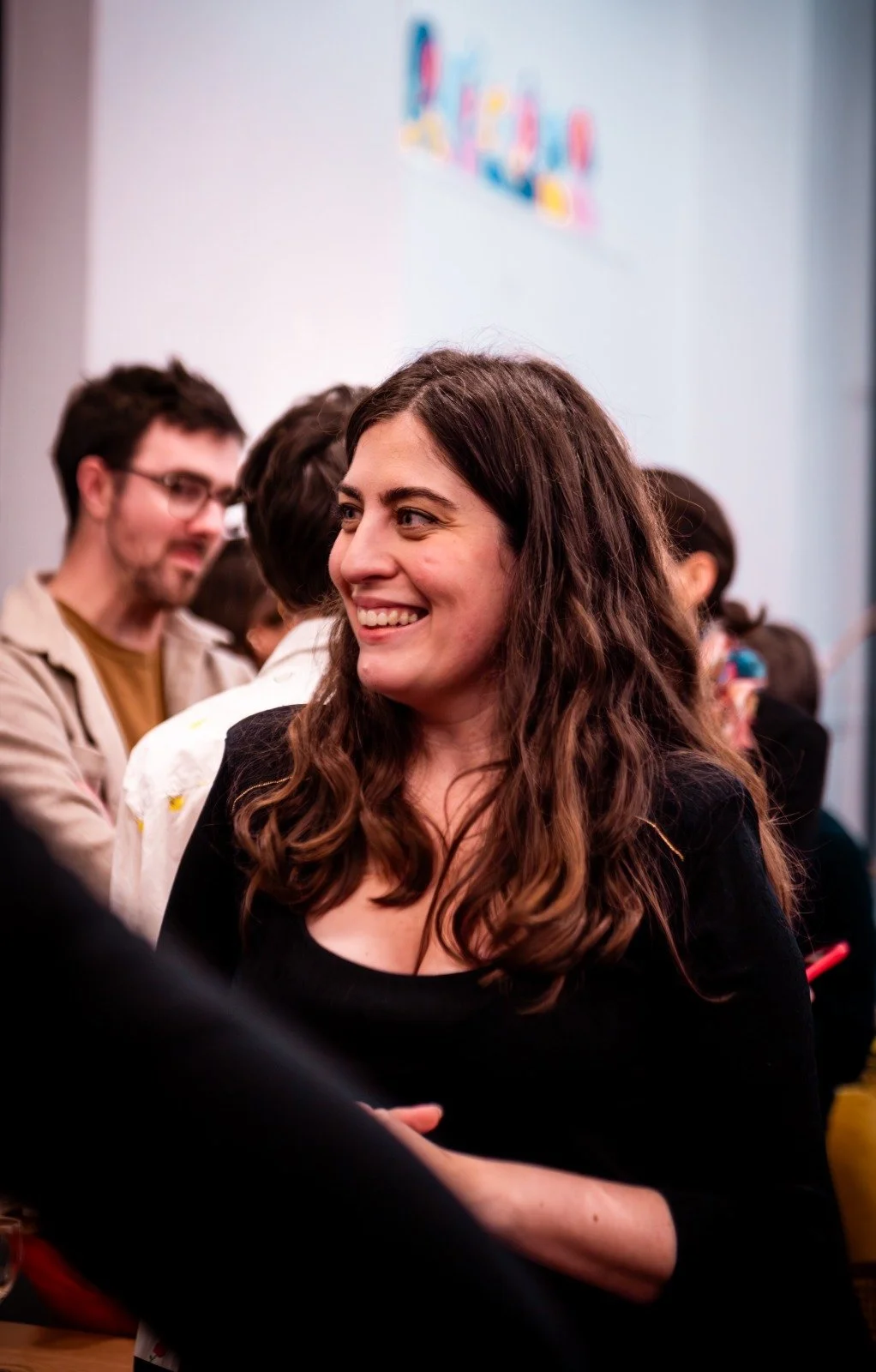 Une femme souriante avec des cheveux longs bruns, entourée de plusieurs personnes lors d'un événement intérieur.