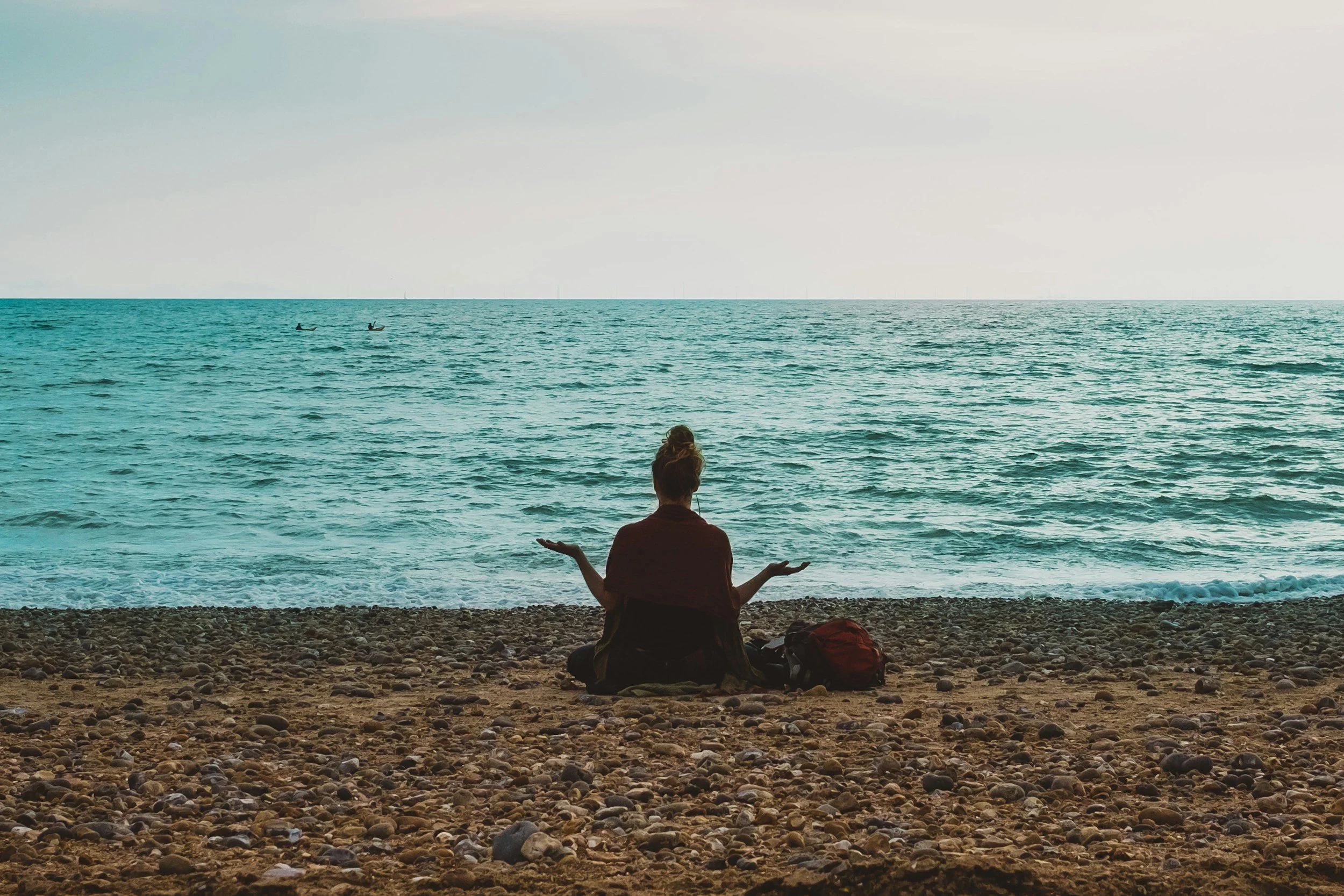 Person sitting on a rocky beach meditating with arms outstretched, facing the ocean with boats in the distance.