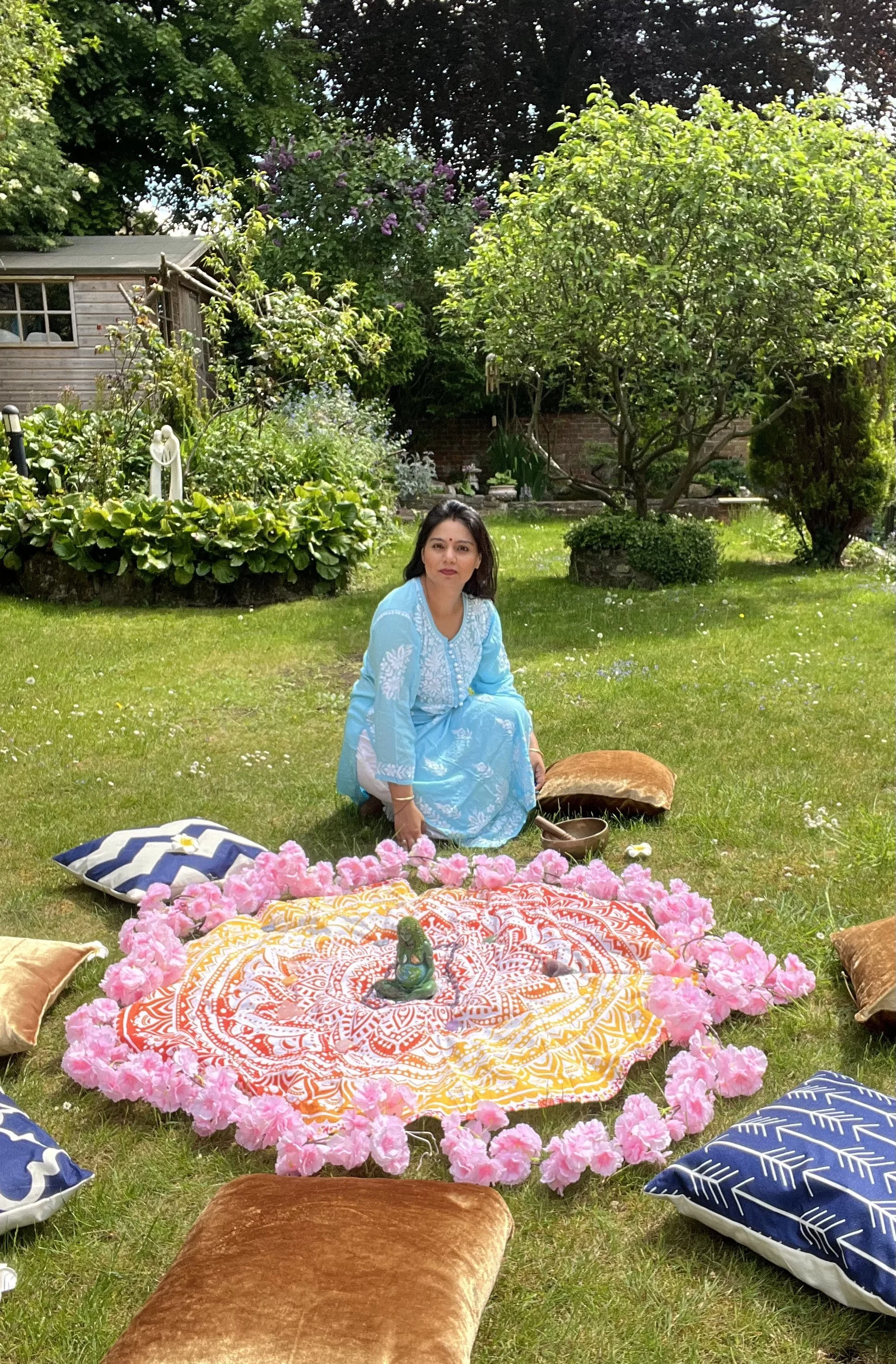 A woman in a blue dress sitting on grass in a garden surrounded by cushions and a decorated cloth with flowers, likely during a cultural or spiritual event.