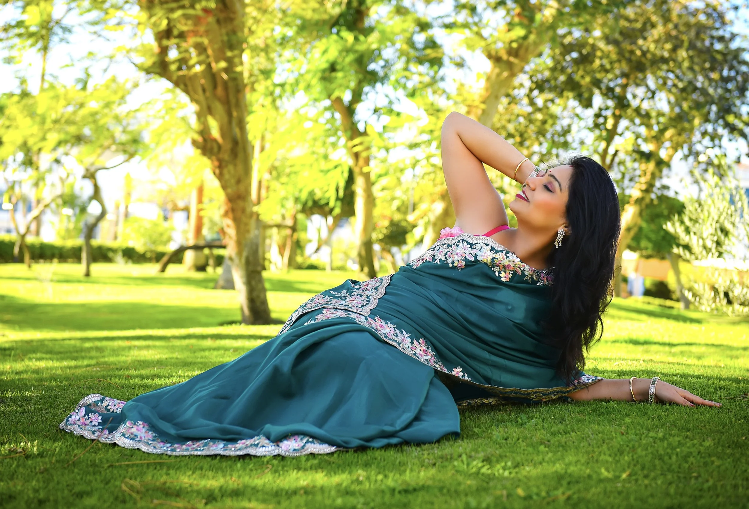 a woman in a teal dress with floral embroidery lying on the grass in a park with trees and sunlight