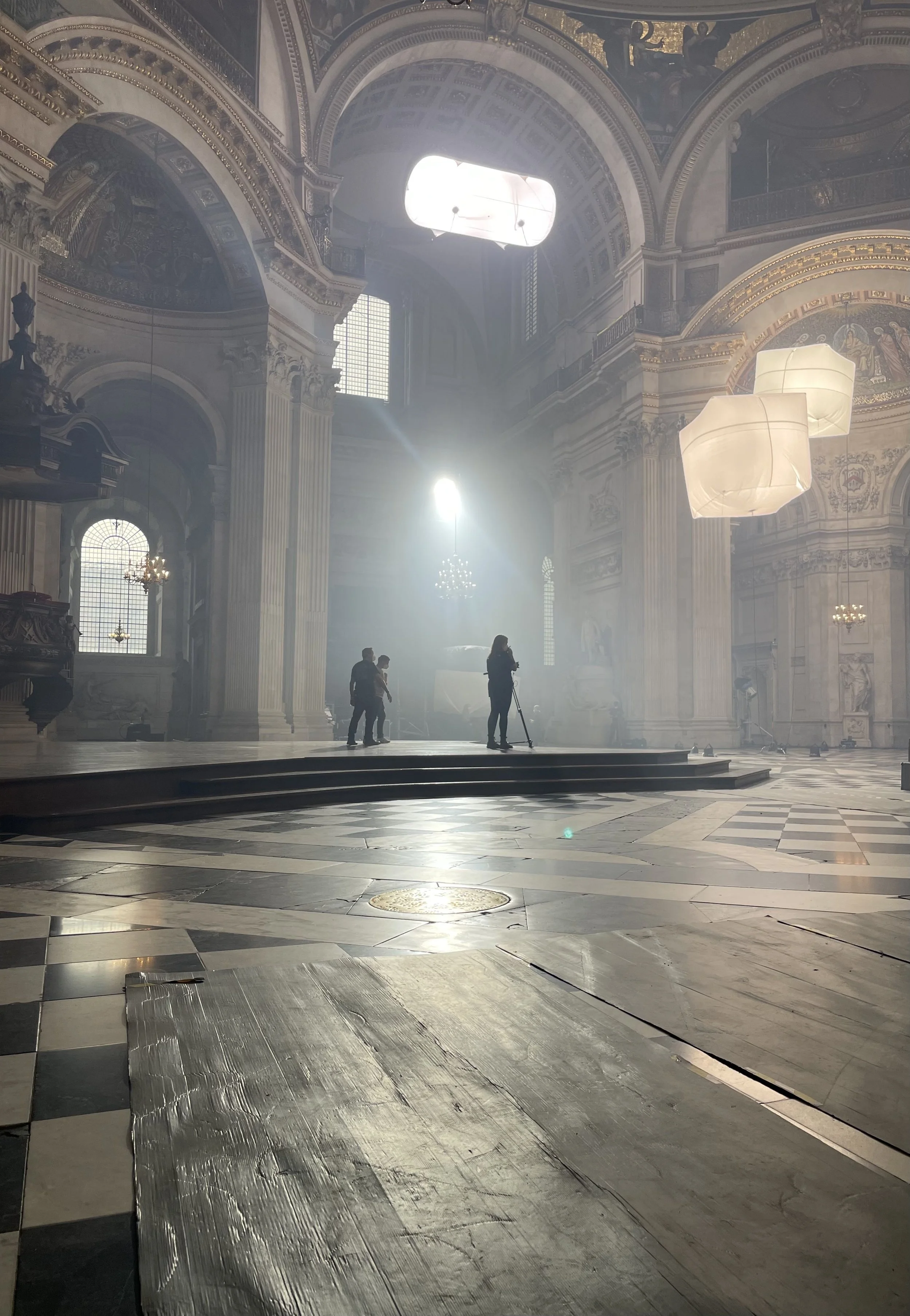 Inside a grand, ornate church or cathedral with high ceilings, large windows, and decorative architecture. Sunlight streams through windows, illuminating a few people standing or walking around, including a woman with a tripod, creating a serene and majestic atmosphere.
