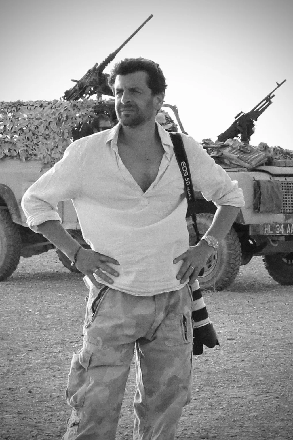 A man standing with hands on hips in front of military vehicles with mounted guns, outdoors during daytime.