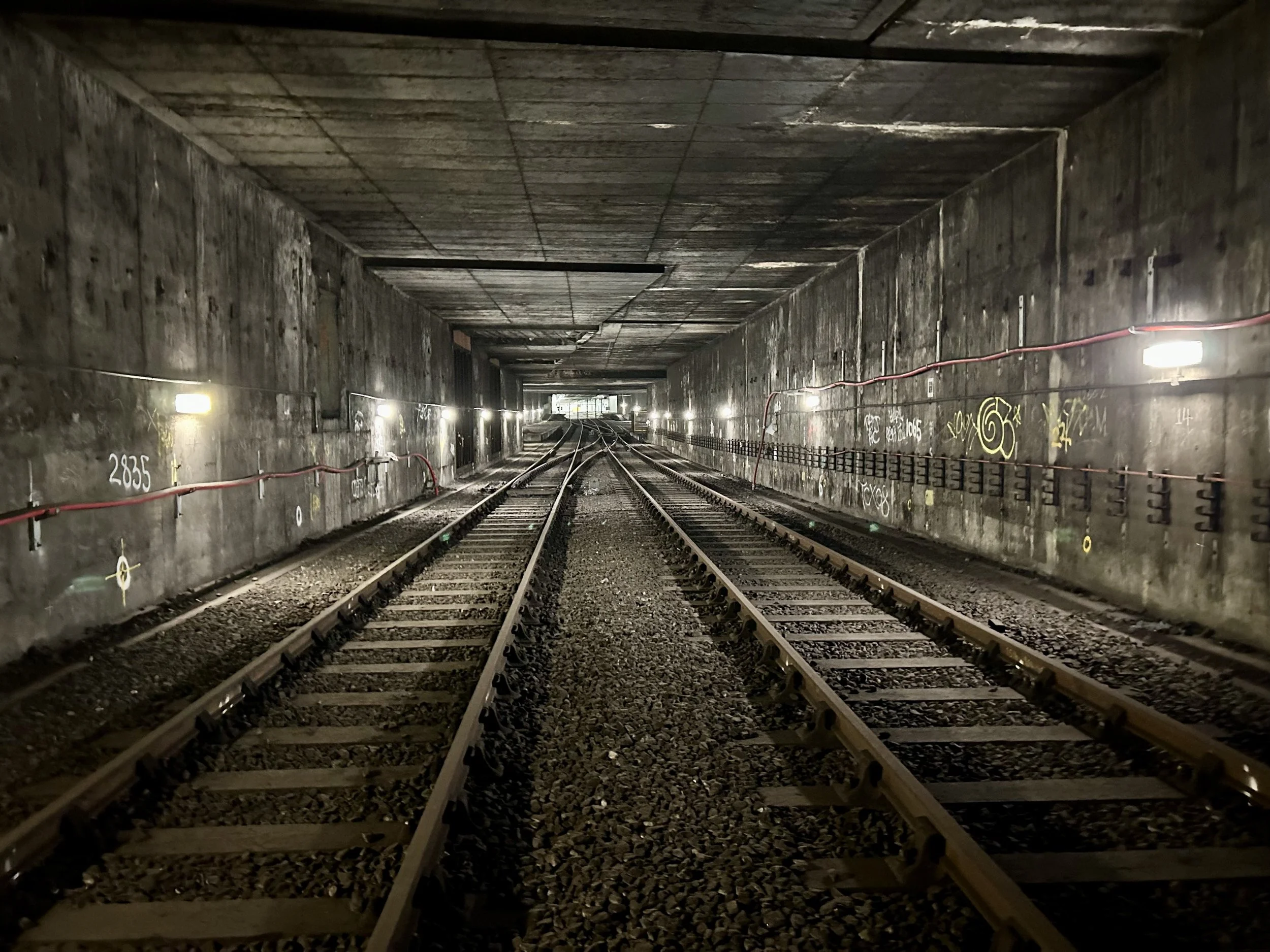 Dark underground train tunnel with railway tracks leading into the distance, illuminated by sparse lights on the walls, with graffiti and safety wires along the sides.