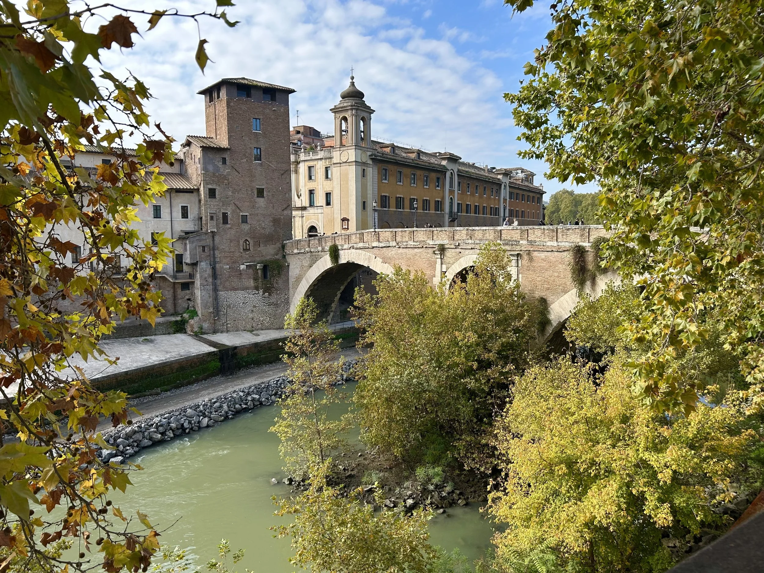 A historic bridge over a river with greenish water, surrounded by trees with autumn foliage, and historical buildings on a bright, partly cloudy day.