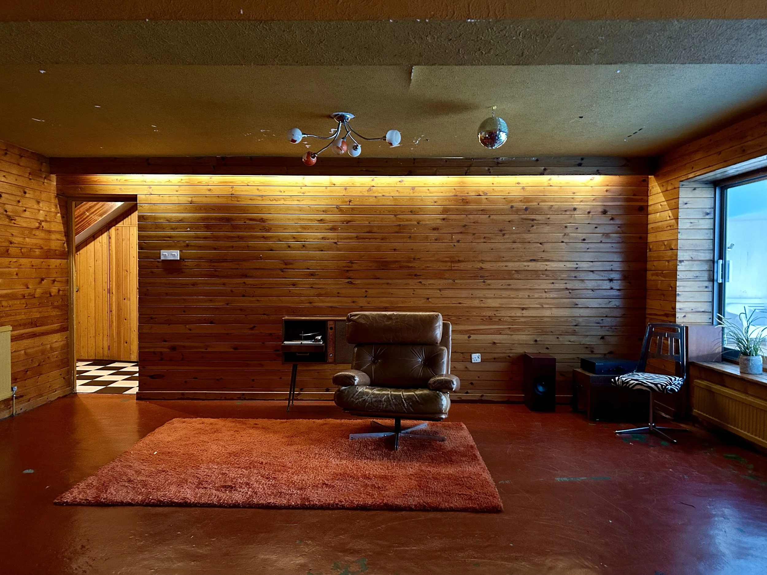A living room with wooden panel walls, a brown leather swivel chair on a red rug, a window with a plant on the windowsill, and a small entertainment setup with speakers. There is a doorway to a checkered black and white floor area and ceiling lights 