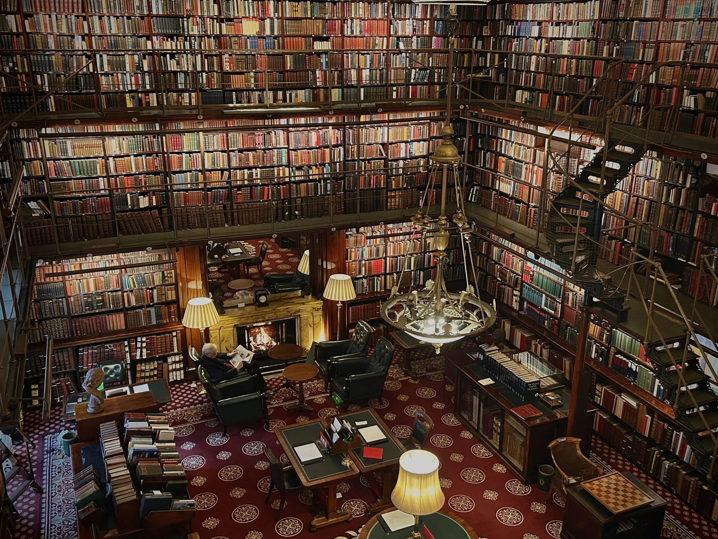 A large, elegant library with multiple floors filled with bookshelves. The ground floor has cozy seating around a fireplace, with a person reading. The bookshelves extend to the ceiling, accessed by spiral staircases, and the room is decorated with v