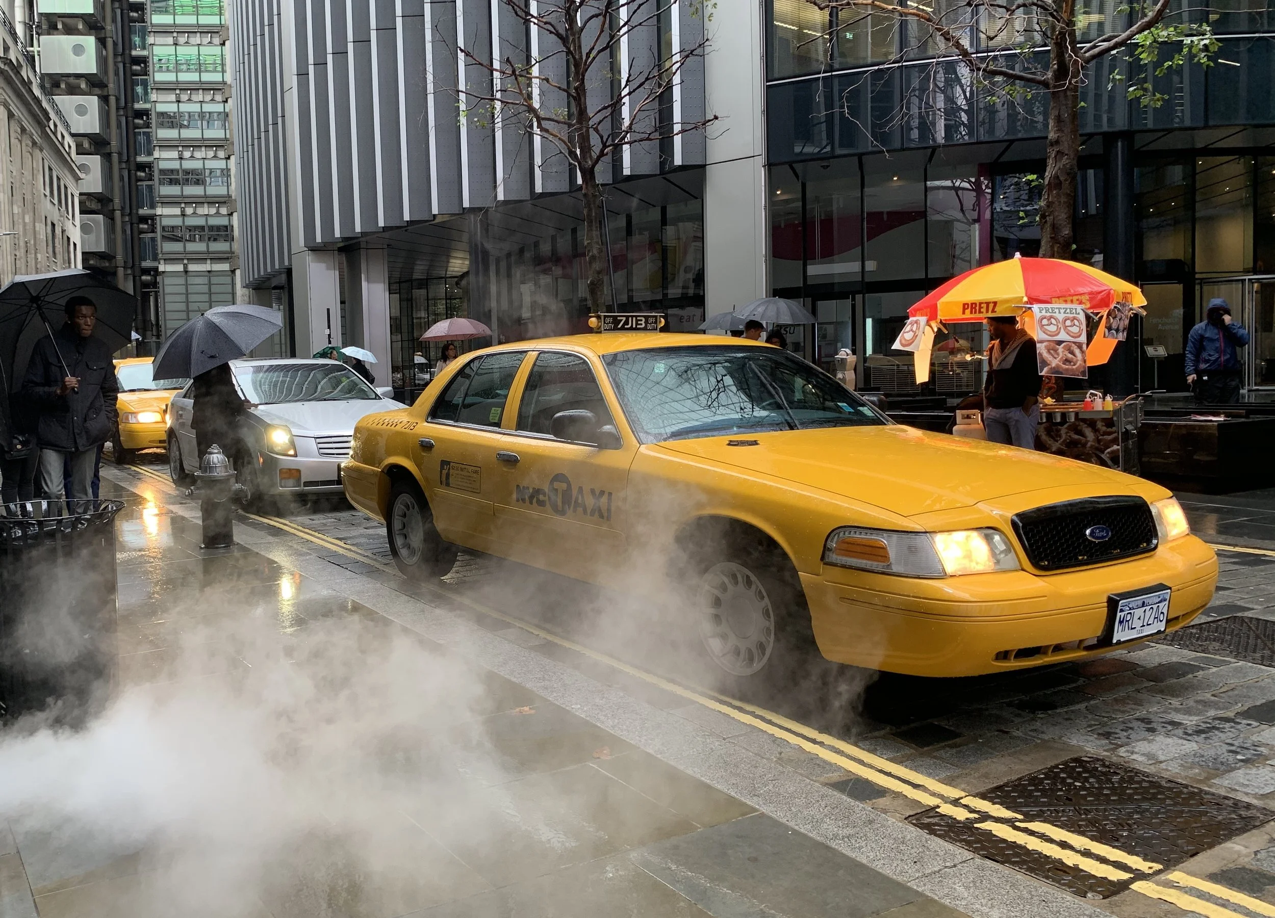 A yellow NYC taxi cab parked on a rainy city street with steam emanating from the ground. Pedestrians with umbrellas walk by, and a street vendor stands under a red and yellow umbrella that reads 'PRETZ'.