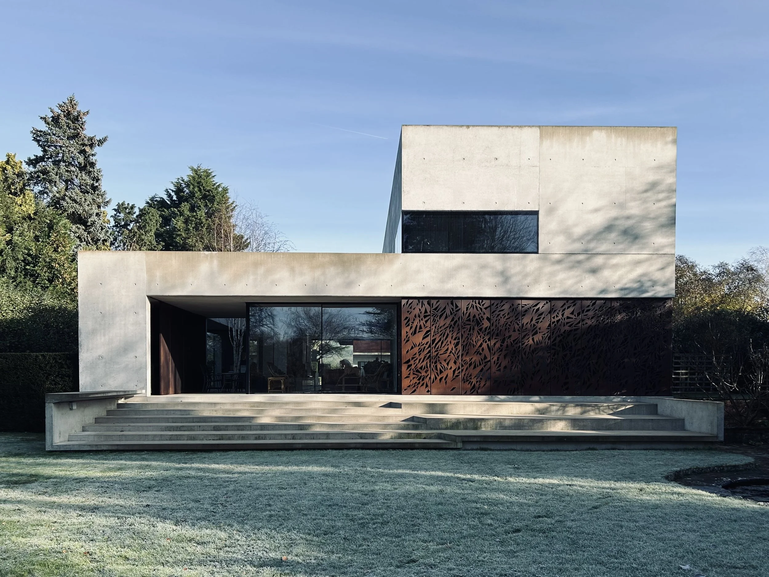 Modern minimalist house with concrete and wood exterior, large glass windows, staircase leading to patio, surrounded by green lawn and trees, under a clear blue sky.