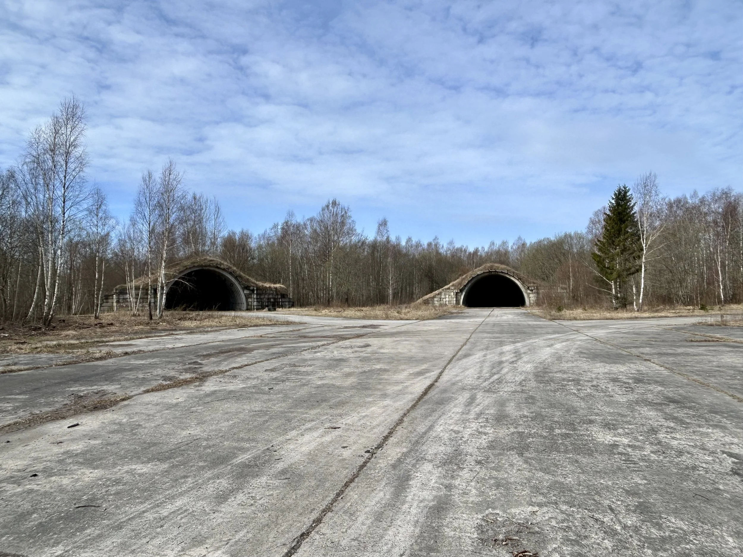 Abandoned concrete road leading into two old, dark train tunnels surrounded by leafless trees and cloudy sky.
