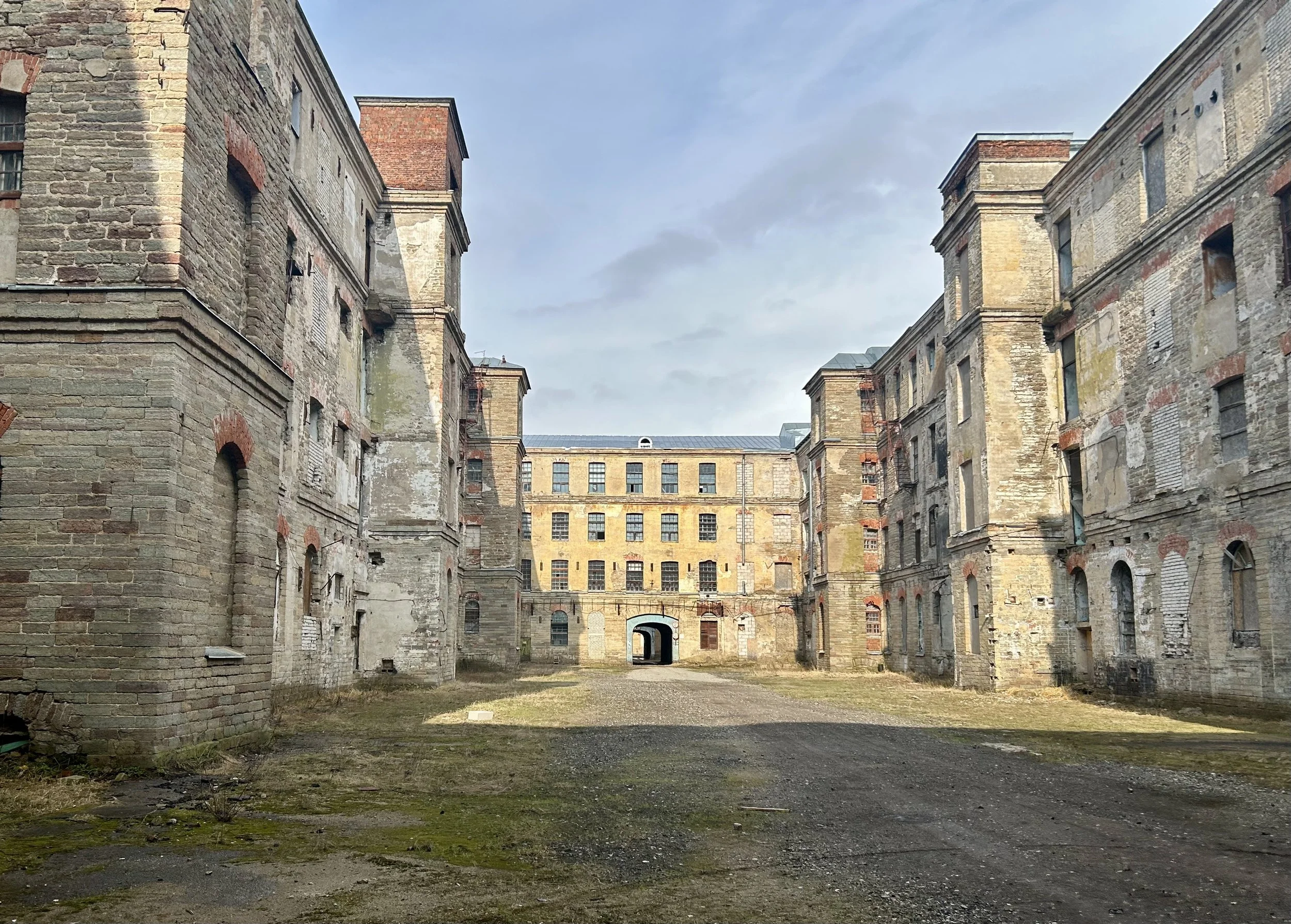 A view of an old, abandoned brick building complex with a central courtyard and a large archway in the background.
