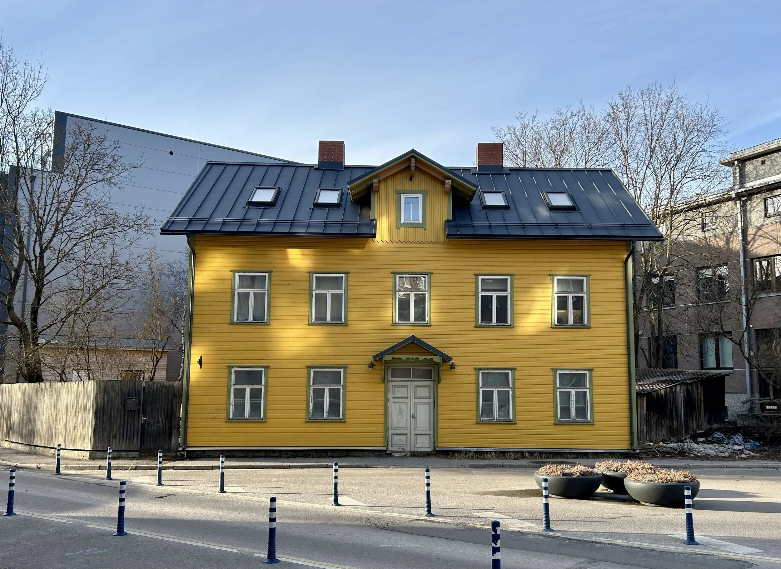 Yellow three-story house with a black metal roof, six windows, and a central door, situated on a city street with potted plants and blue-striped traffic posts in the foreground.