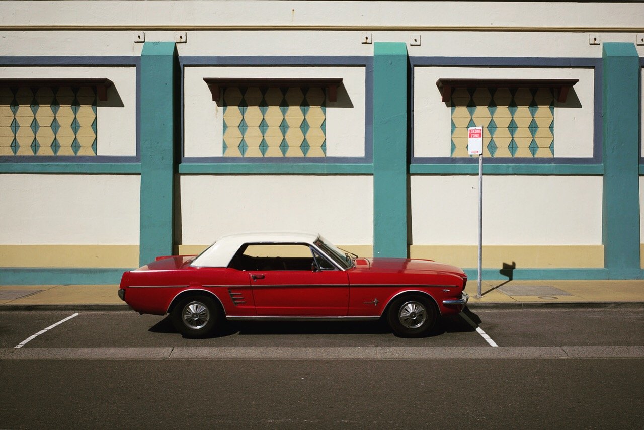 A classic red convertible car with a white top parked in a space on the street against a colorful building with teal accents and patterned windows.