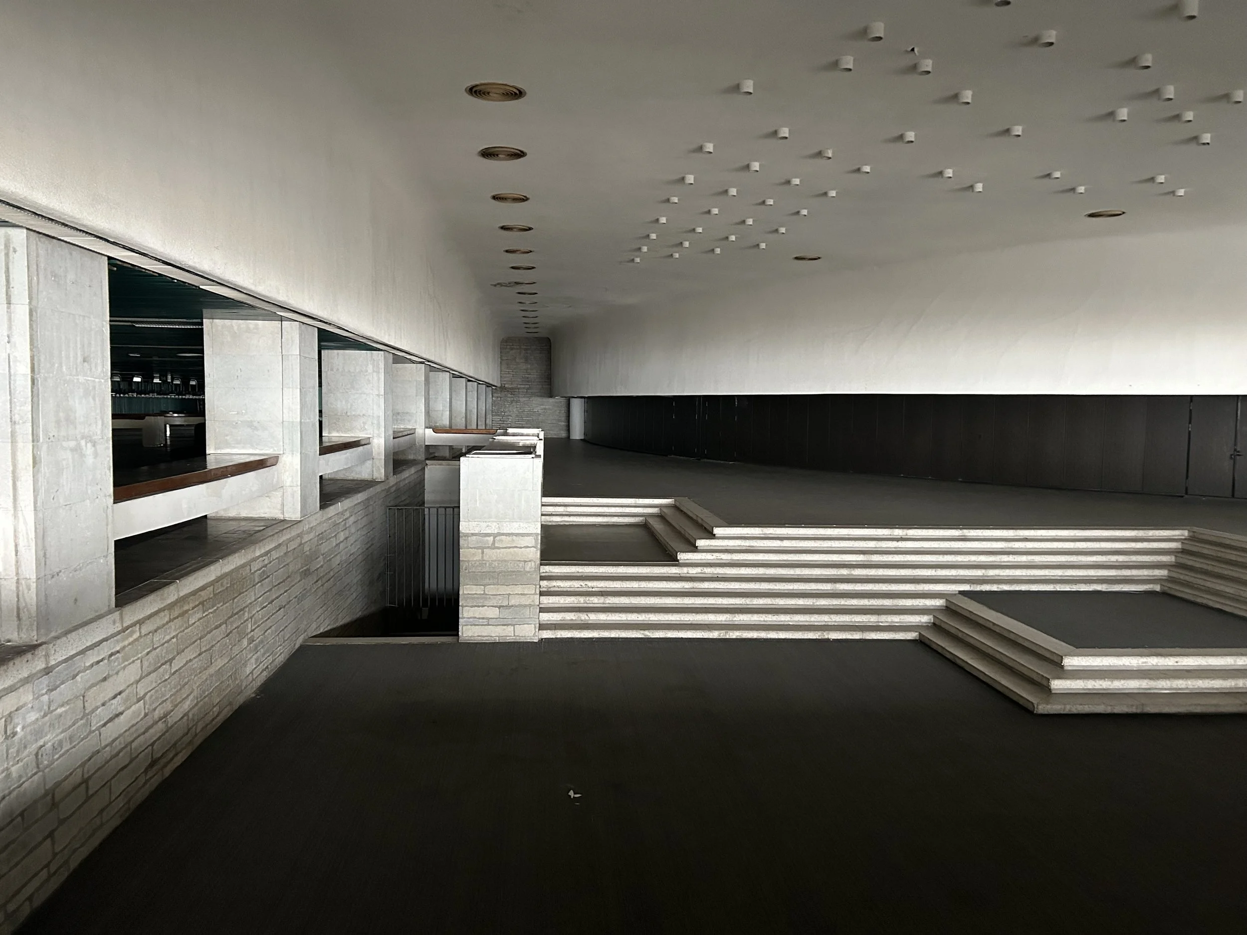 Empty modern interior space with stairs, concrete and brick walls, and a white ceiling with numerous small lights.