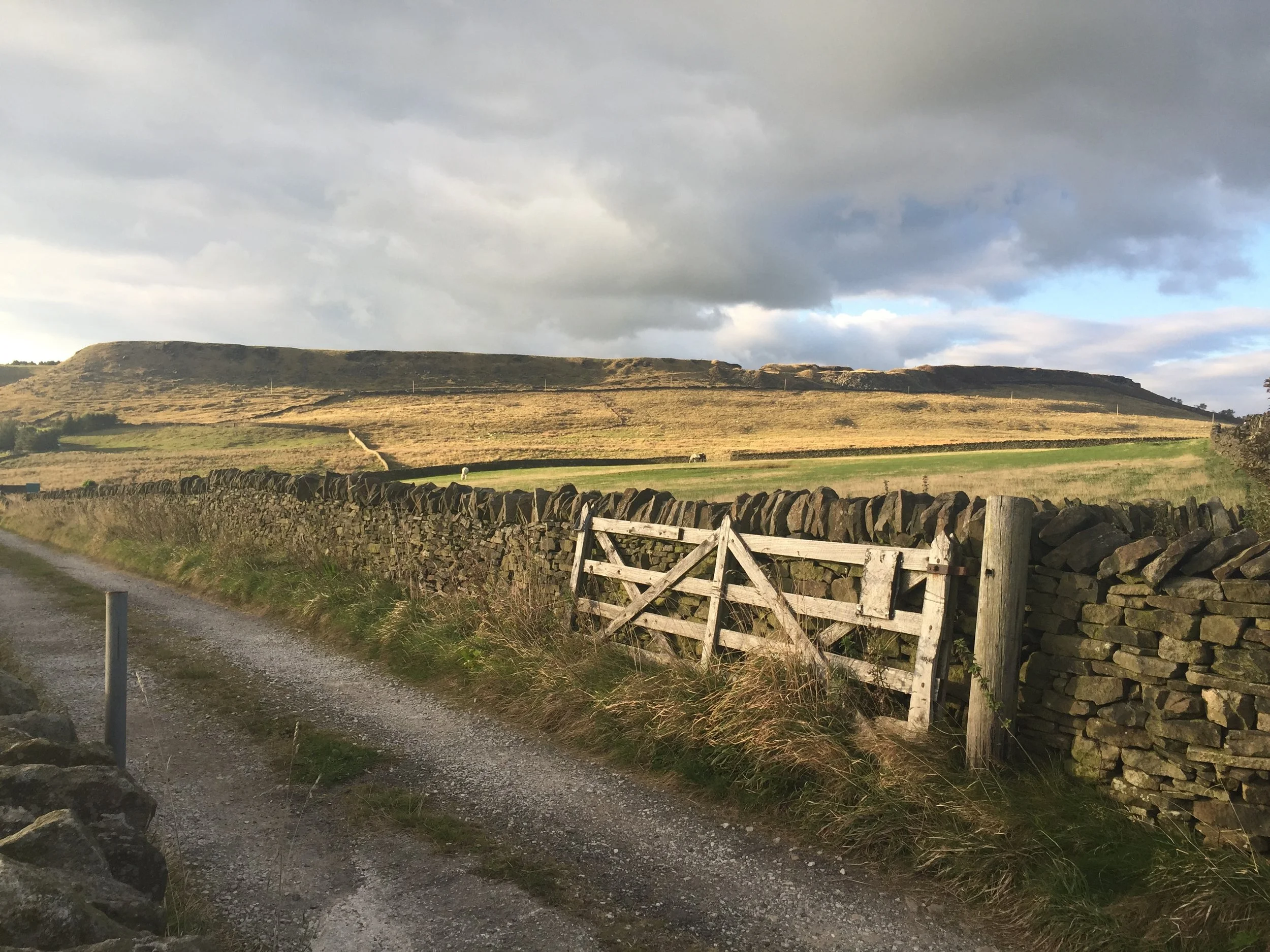 A rural landscape with a gravel road, a stone wall, and a weathered wooden gate in the foreground. Rolling hills extend into the distance under a cloudy sky.