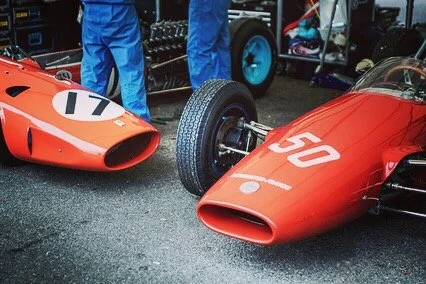 Two vintage red race cars with aerodynamic designs and large air intakes, parked outdoors with people around.