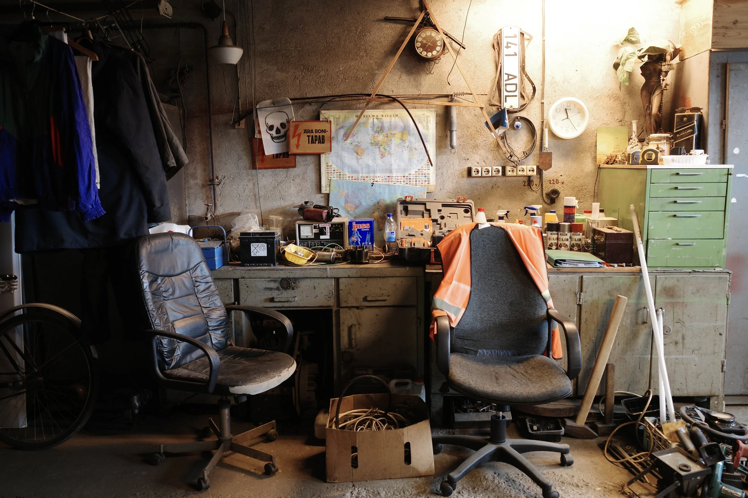 A cluttered workspace with two office chairs, a workbench with various tools and equipment, a green cabinet, and miscellaneous items. The wall has a map, a skull sticker, and a clock, all illuminated by a warm light.