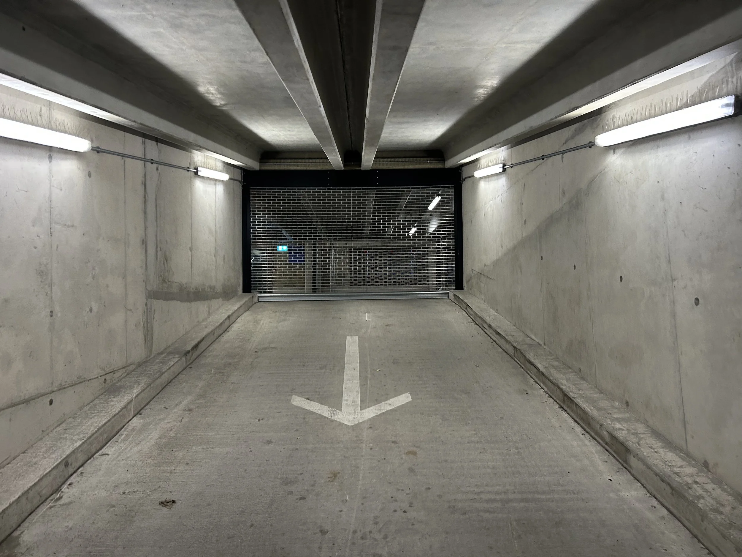 Underground parking garage entrance with a downward arrow painted on the concrete floor, concrete walls, and several fluorescent lights on the ceiling.