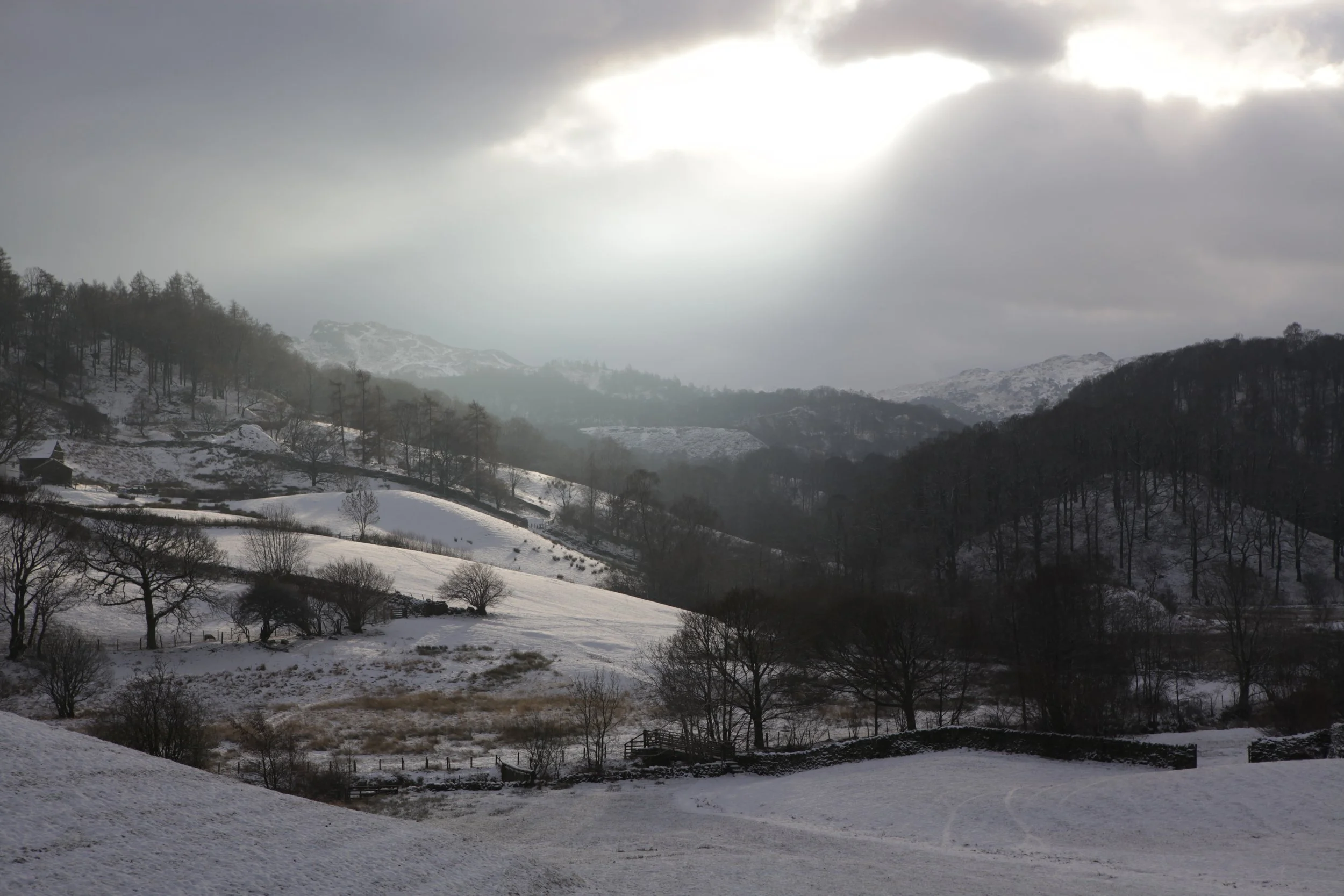 Snow-covered hills and mountains with sparse trees, under a cloudy sky and some sunlight breaking through.