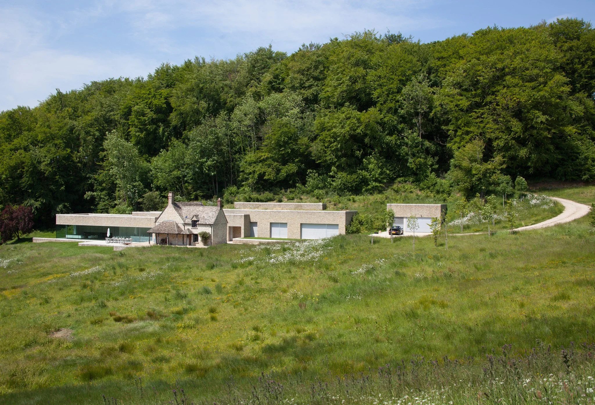 Modern house with stone walls and glass windows on a green hillside, surrounded by trees under a blue sky.