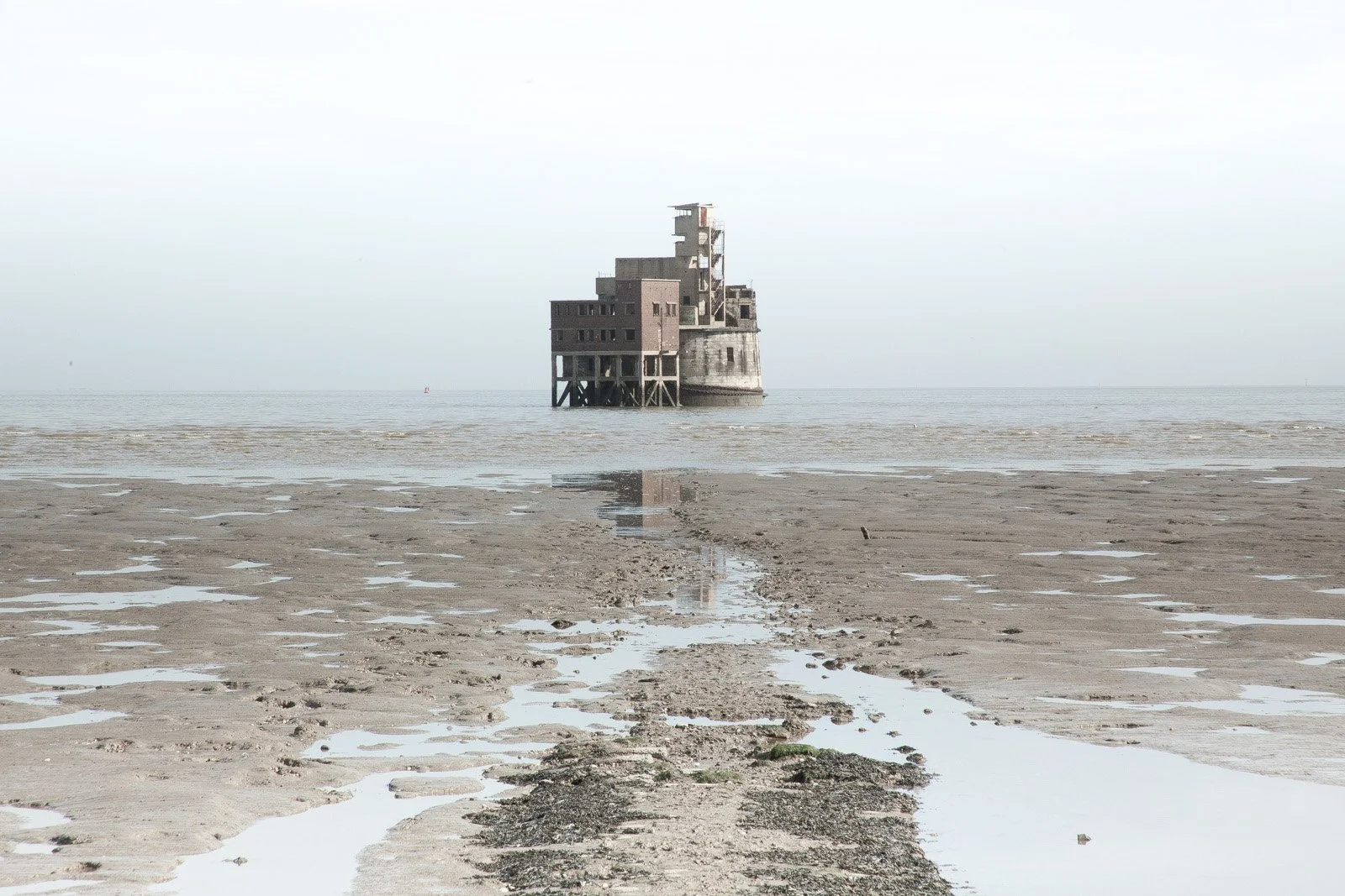 A building on stilts in the ocean, viewed from the shore with muddy, rippled sand and shallow water, under a cloudy sky.