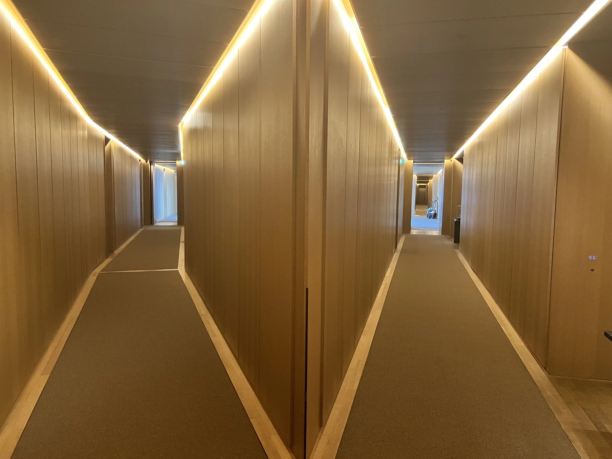 Side-by-side comparison of two hallway images with wooden walls and ceiling, soft lighting along the edges, and brown carpeting, leading to different rooms or areas at the end.