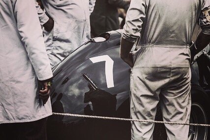 Close-up of a dark-colored car part with the number 7 on it, surrounded by people in gray coats or overalls.
