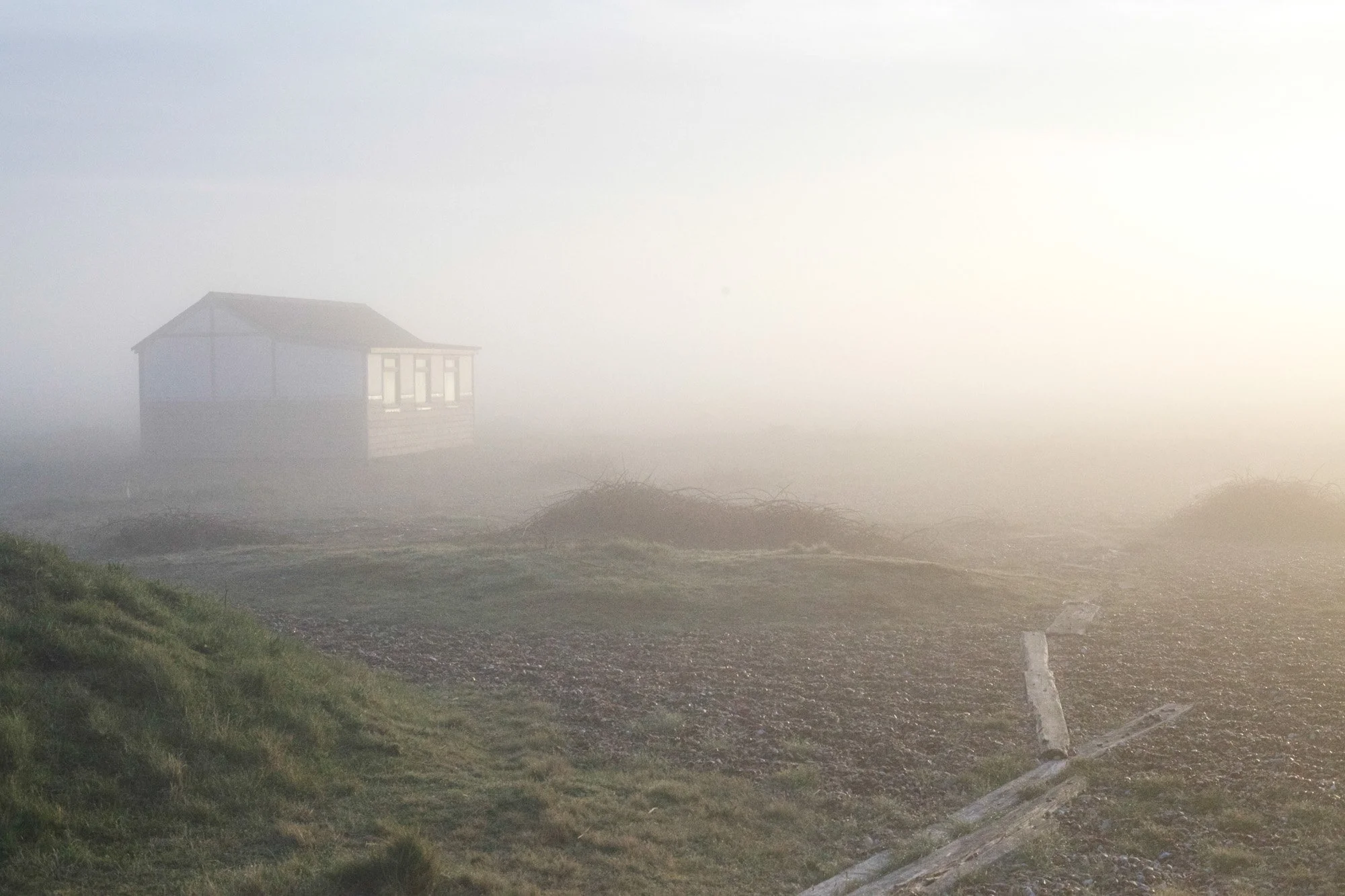 A foggy outdoor scene with a small house in the background, partially obscured by fog. The foreground has a grassy area and a stone pathway.
