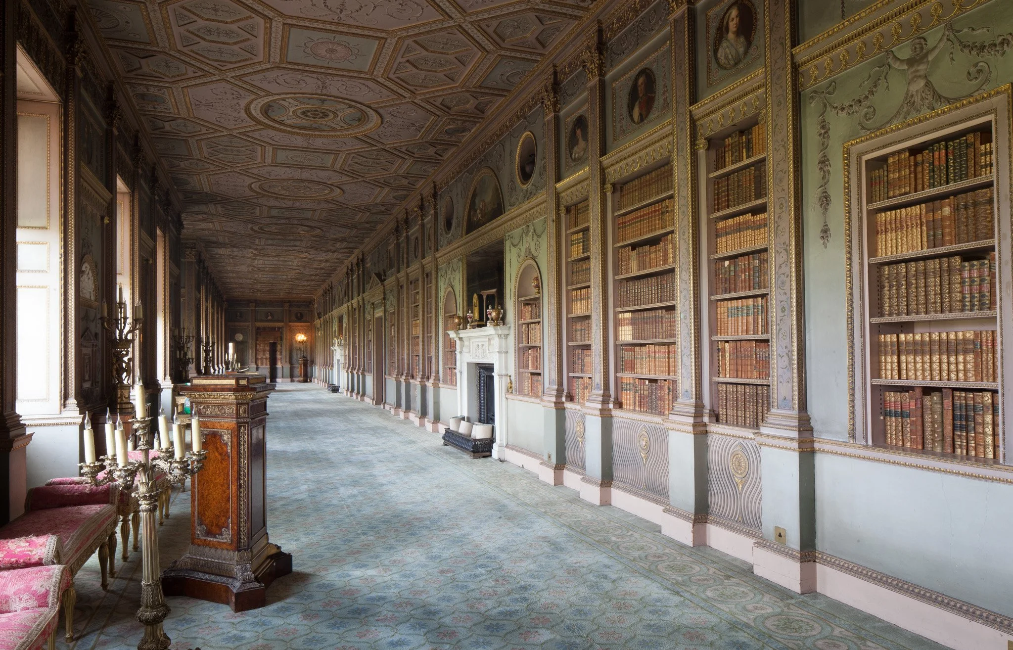 Long, ornate library hallway with tall wooden bookshelves filled with leather-bound books, a marble fireplace, and vintage furniture. Sunlight streams in through large windows.