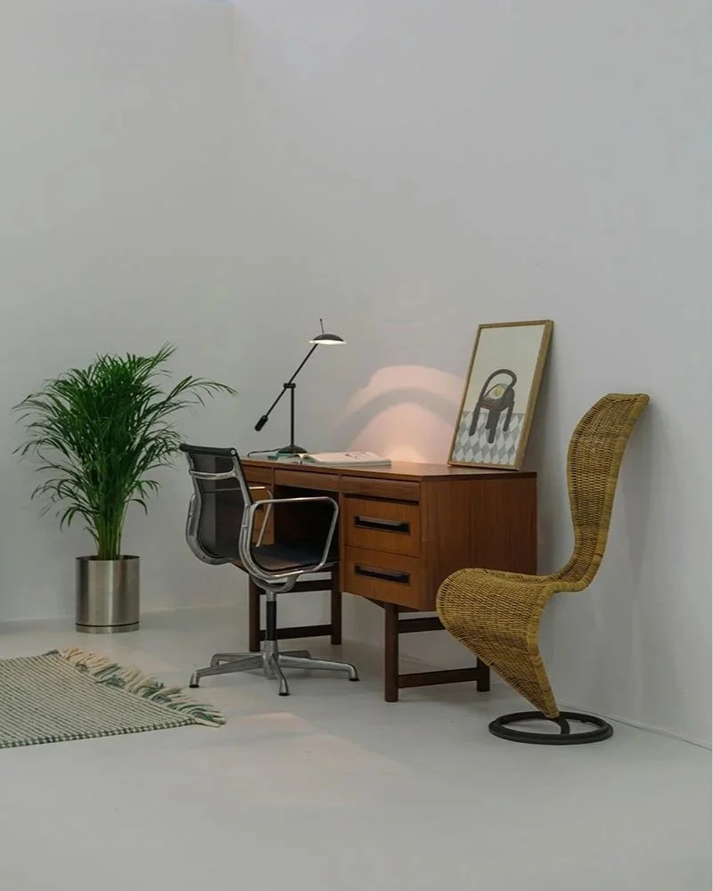 Un bureau en bois avec une lampe, une plante verte en pot, une chaise de bureau, une chaise en rotin, et un tableau posé contre le mur blanc.