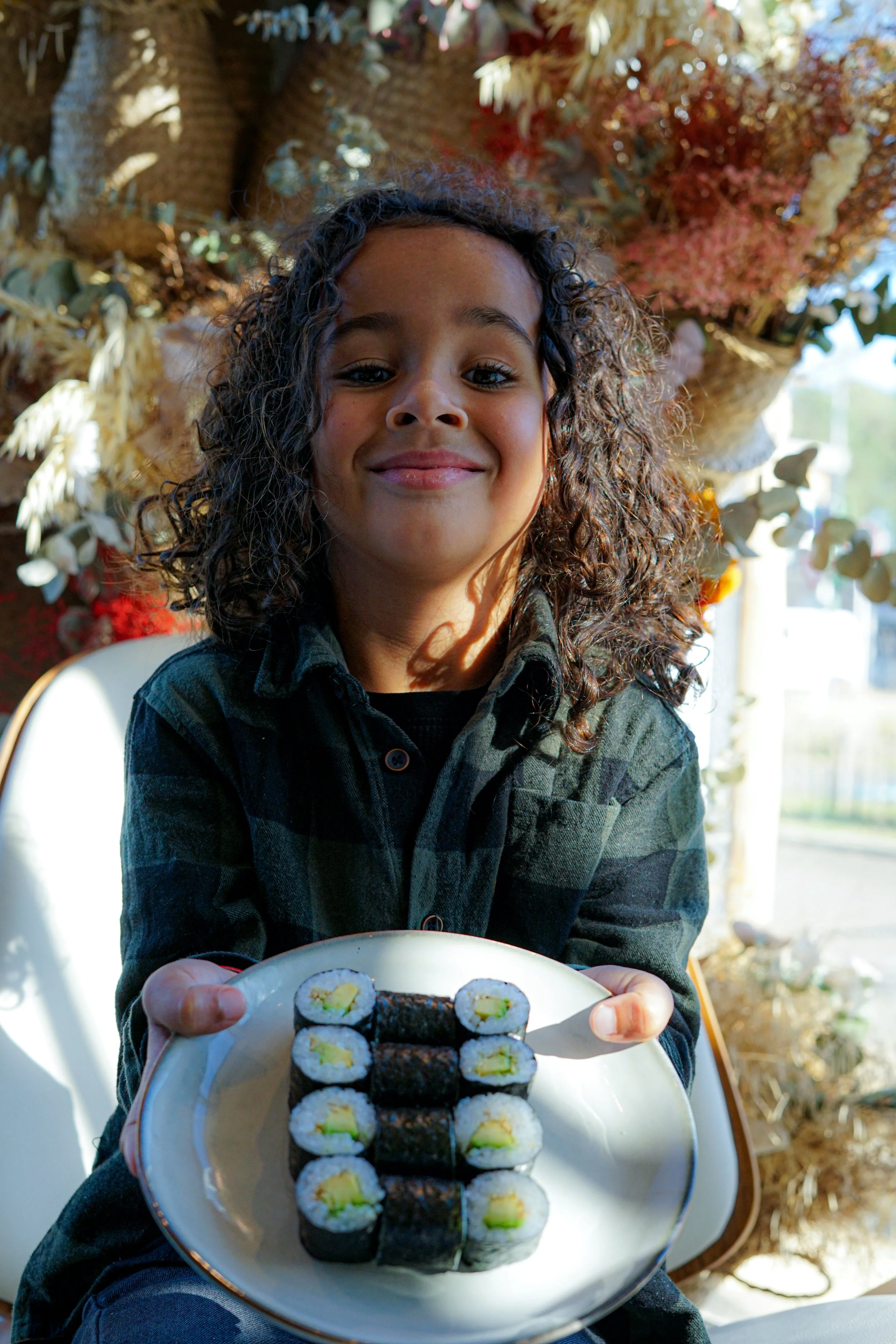 Une jeune fille avec des cheveux bouclés tient un plateau de sushi et sourit. Les sushis sont disposés en rangée sur une assiette. En arrière-plan, il y a des décorations florales ou végétales et une lumière naturelle ambiance chaleureuse.